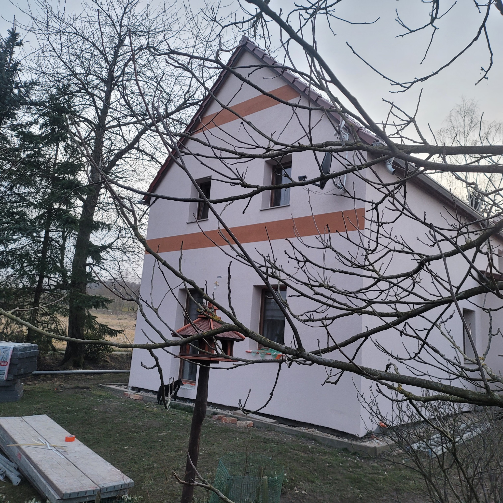 Otynkowany dom jednorodzinny z brązowym pasem dekoracyjnym, widoczny zza gałęzi drzew. Na trawniku leżą materiały budowlane i stoi karmnik dla ptaków.