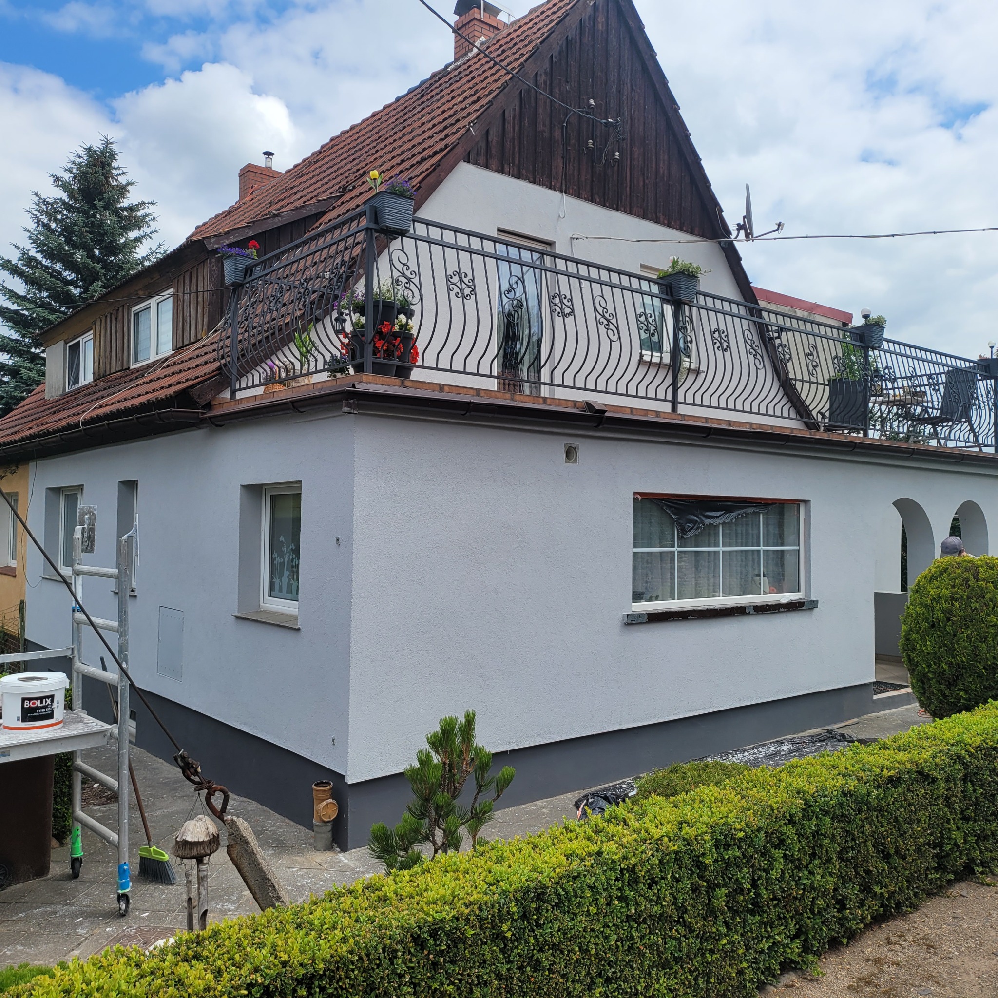 Odnowiona elewacja domu z balkonem, świeżo po malowaniu na szaro. Widoczne rusztowanie, puszka farby Bolix i starannie przycięty żywopłot.