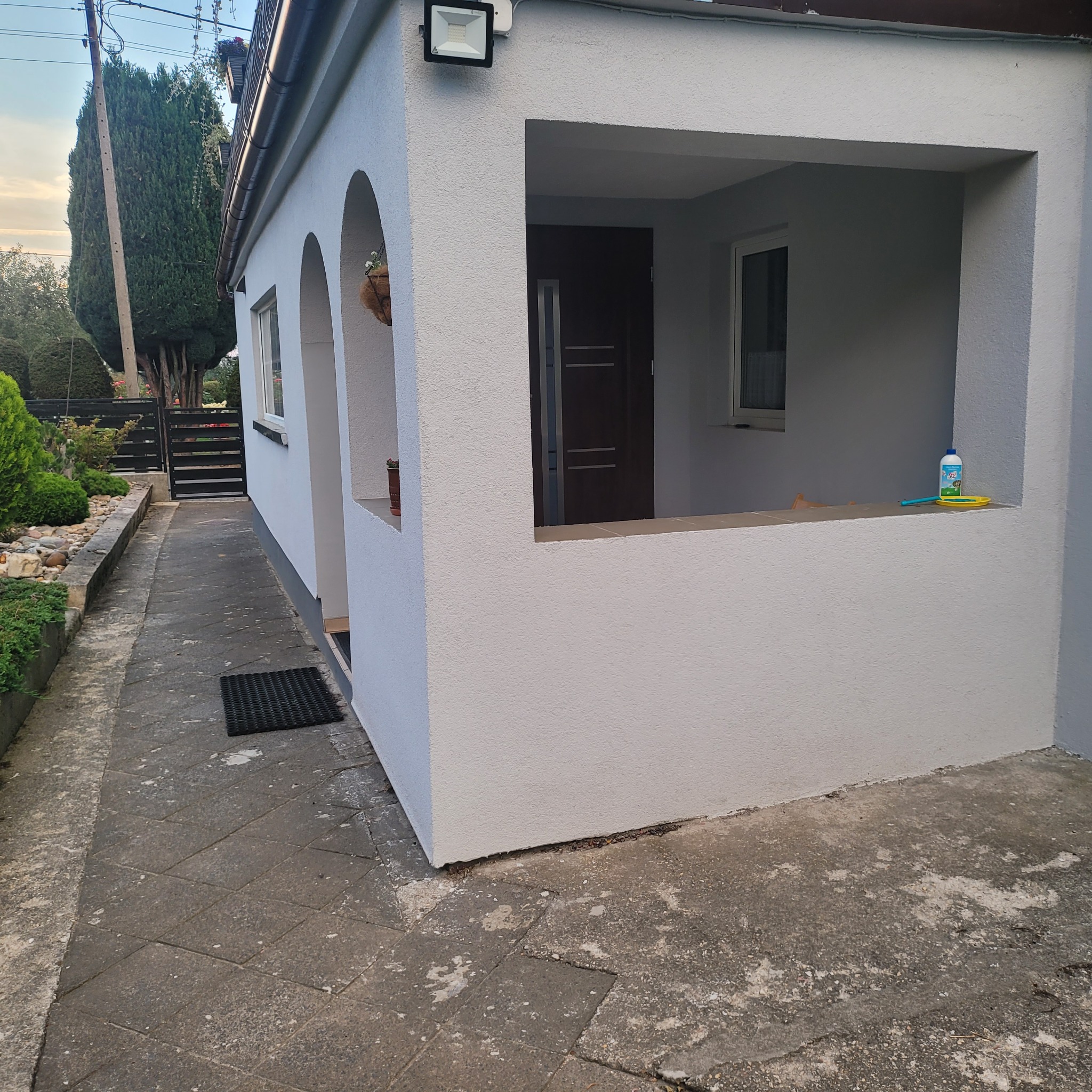 Odnowiona elewacja domu z łukowym wejściem i otwartym gankiem. Świeża, jasnoszara farba, widoczna boczna ścieżka i zieleń w tle. Na ganku butelka płynu i zabawki.