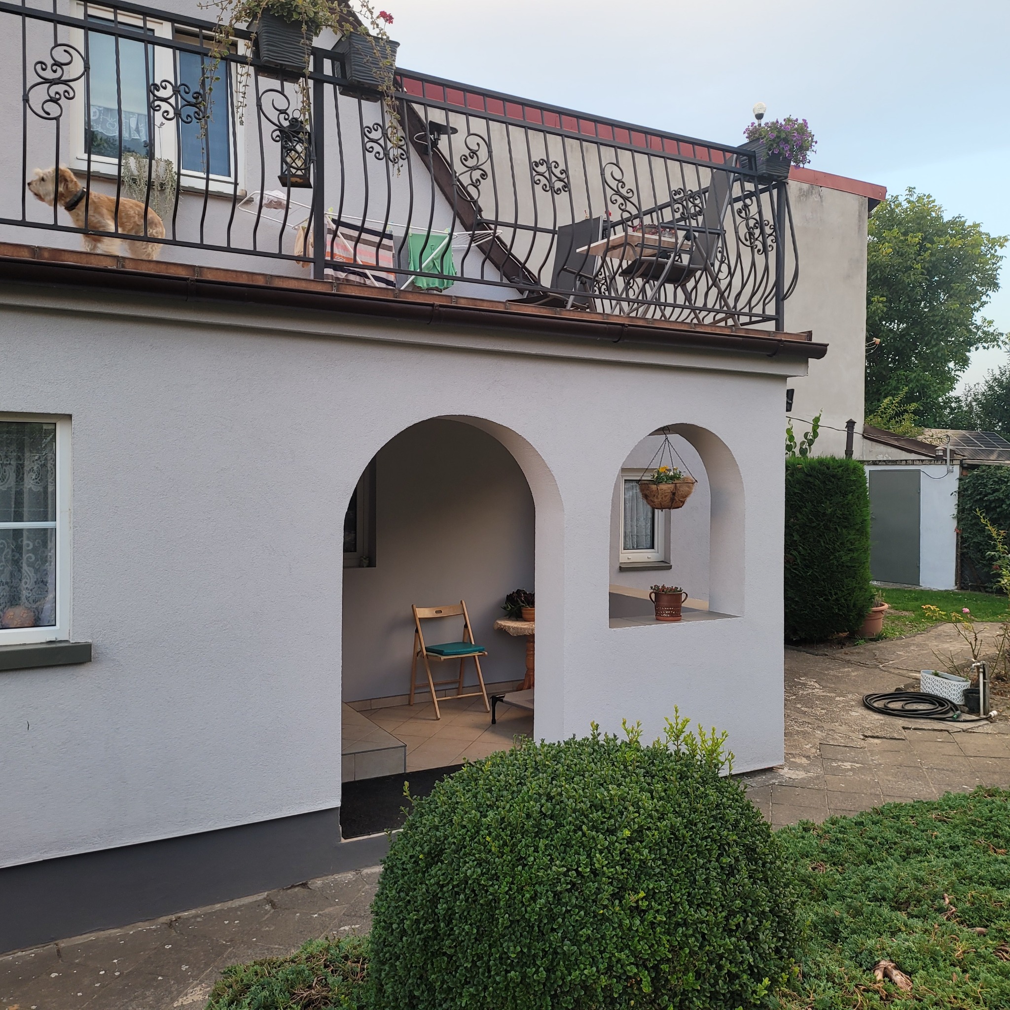 Biały budynek z balkonem z kutą balustradą, na którym stoi pies. Na parterze arkady z krzesłem i stolikiem. W oknach firanki. W tle zieleń.