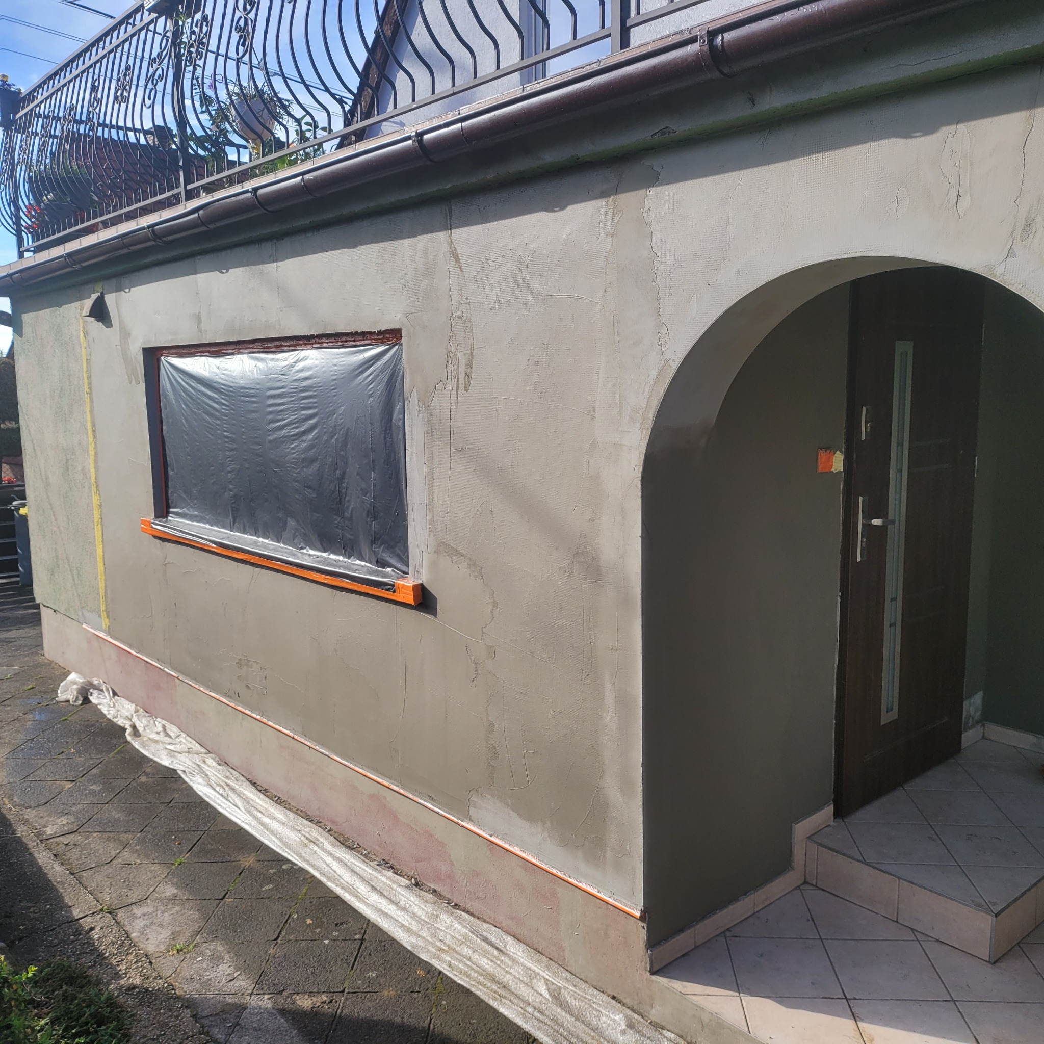 Ściana budynku w Biedrzychowicach po nałożeniu tynku, okno zabezpieczone folią. Widoczny fragment elewacji z drzwiami wejściowymi i balkonem na górze.