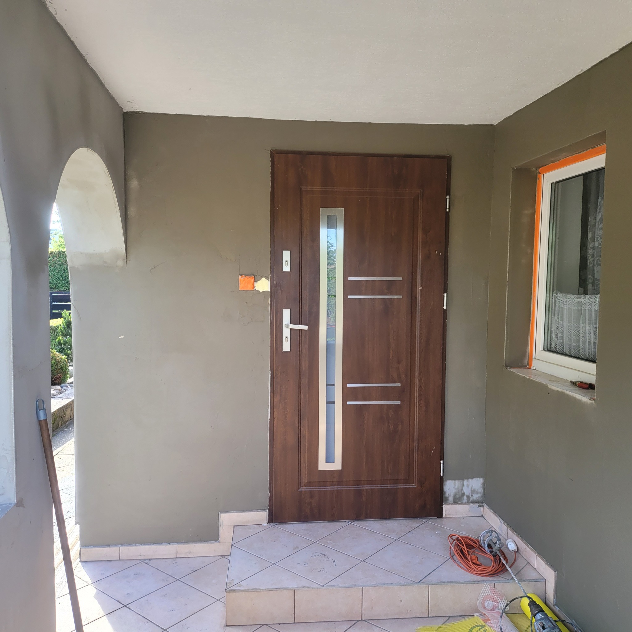 Montaż brązowych drzwi wejściowych z szybą na tle szarej elewacji w trakcie prac wykończeniowych. Widoczne narzędzia i fragment okna z firanką.