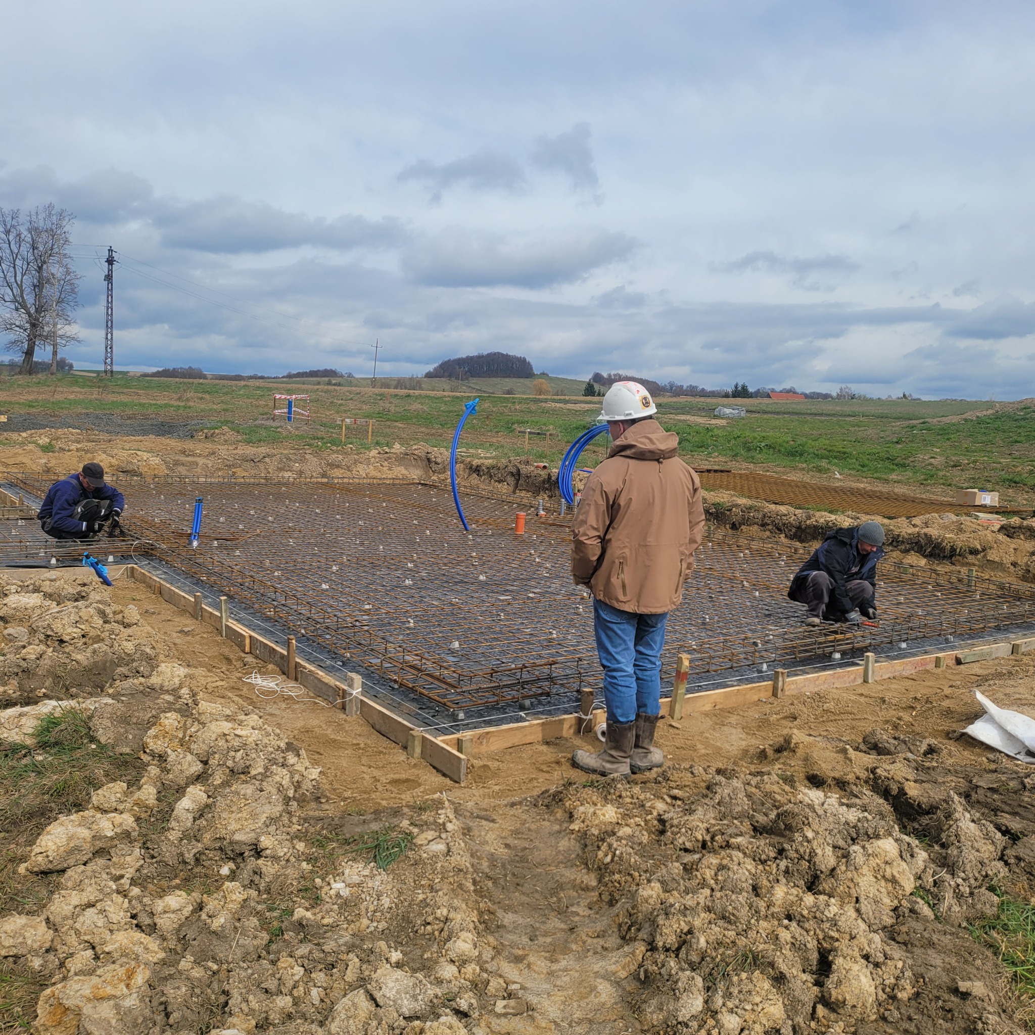 Zbrojenie fundamentów pod budynek jednorodzinny. Widoczne szalunki, stal zbrojeniowa, rury instalacyjne i pracujący robotnicy w kaskach. Tło: pole i pochmurne niebo.