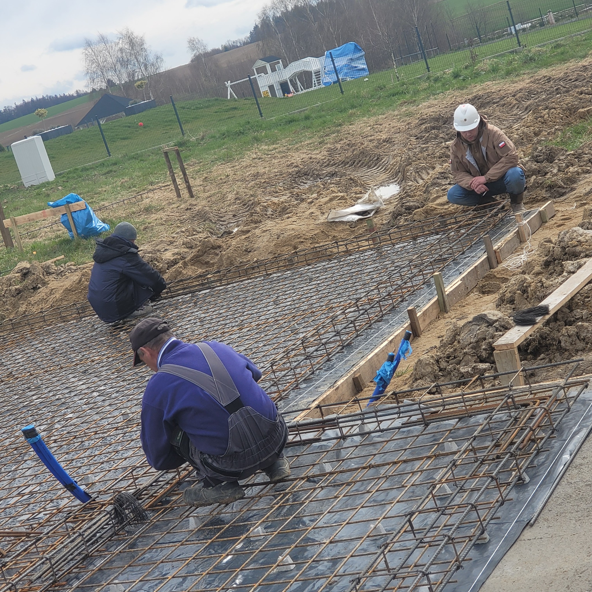 Zbrojenie fundamentów pod budowę domu jednorodzinnego. Widoczni pracownicy w trakcie montażu siatki zbrojeniowej na folii. W tle krajobraz wiejski.