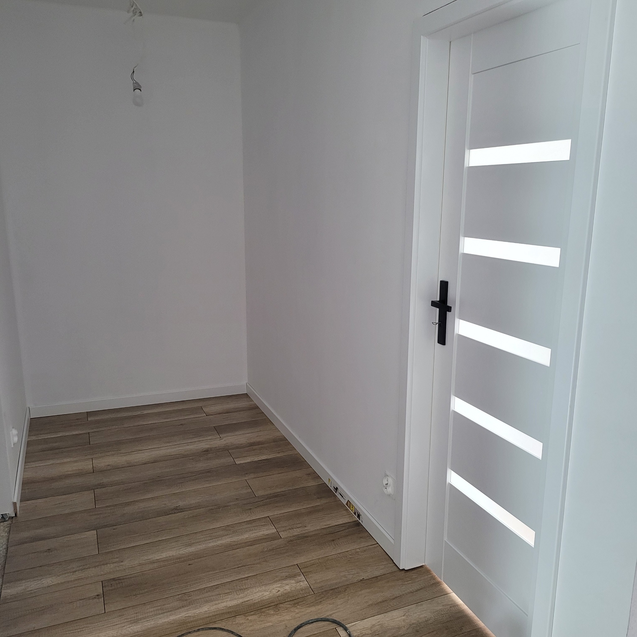 Wnętrze po remoncie: drewnopodobne płytki na podłodze, białe ściany i nowoczesne białe drzwi z przeszkleniami. Widoczne gniazdko elektryczne.