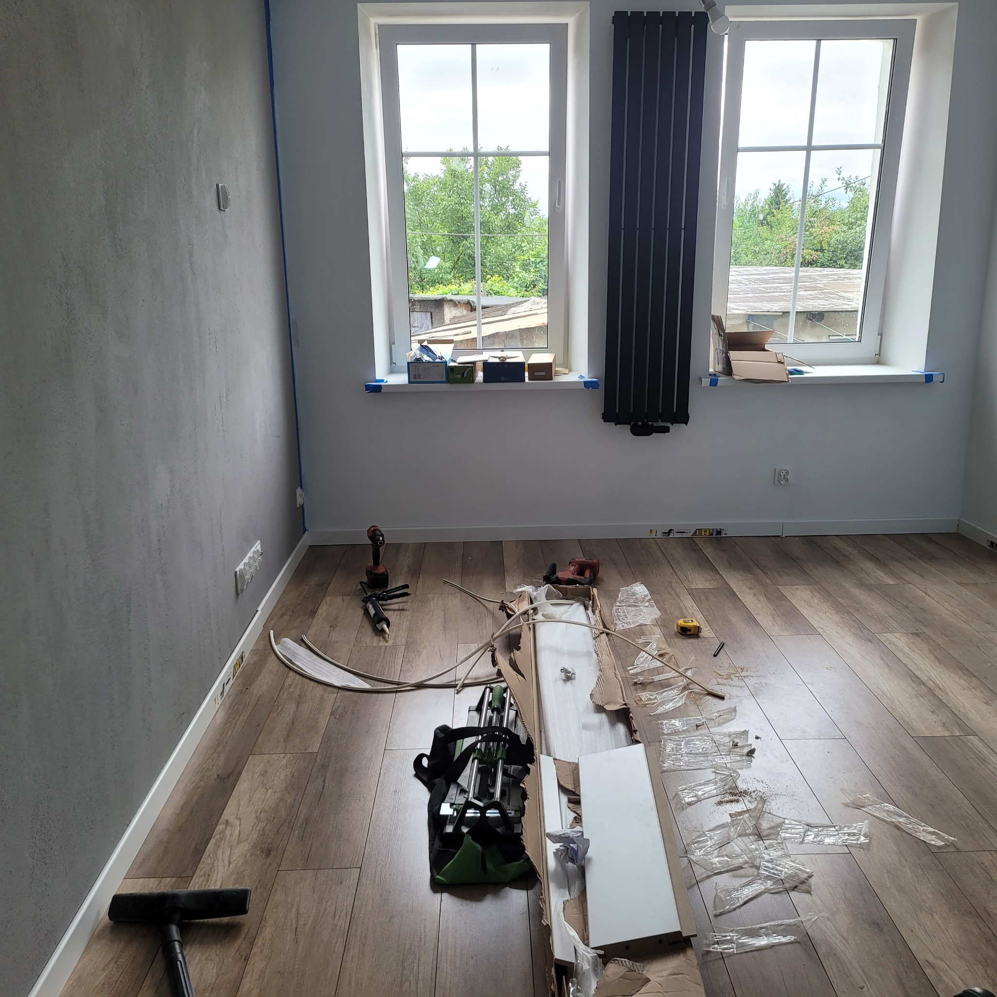 Wnętrze pokoju w trakcie wykańczania: panele podłogowe, narzędzia glazurnicze, listwy przypodłogowe, okna z widokiem na zieleń. Szara ściana z widoczną instalacją elektryczną.