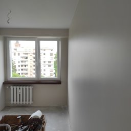 Kacper Golba - Pokój po malowaniu ścian na jasny odcień, widoczne narzędzia malarskie w drewnianej skrzynce, grzejnik pod oknem z widokiem na budynki mieszkalne.