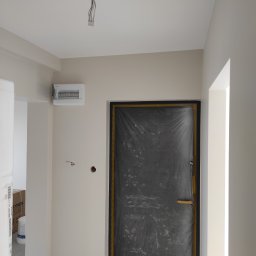 Kacper Golba - Przygotowane do malowania wnętrze: zabezpieczone folią drzwi, pomalowane ściany na jasny kolor, widoczna skrzynka elektryczna i wystające kable z sufitu.