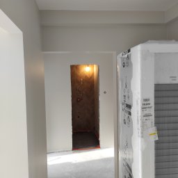 Kacper Golba - Wnętrze w trakcie prac, widoczna ściana pomalowana na szaro, w tle otwór drzwiowy do wykończonej łazienki z brązowymi płytkami ułożonymi w jodełkę, na pierwszym planie nowy sprzęt AGD w folii...