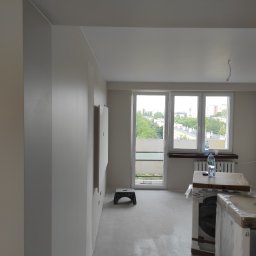 Kacper Golba - Wnętrze w trakcie prac wykończeniowych: świeżo pomalowane na szaro ściany, okno z widokiem na zieleń, zapakowane sprzęty AGD i puszka z farbą na podłodze.