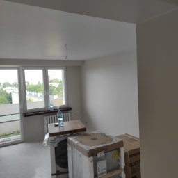 Kacper Golba - Puste, jasne wnętrze w trakcie przygotowań do instalacji sprzętu AGD, z zapakowanymi pralkami i suszarkami oraz butelkami wody na parapecie okiennym.