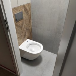 Kacper Golba - Wisząca toaleta w nowoczesnej łazience z szaro-brązowymi płytkami, widoczny fragment podłogi z parkietu i rama drzwi.