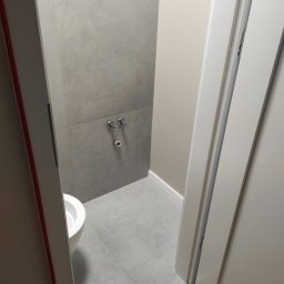 Kacper Golba - Fragment łazienki z szarymi płytkami, widoczna muszla klozetowa, podejścia wodne, białe listwy przypodłogowe i fragment otwartych białych drzwi.