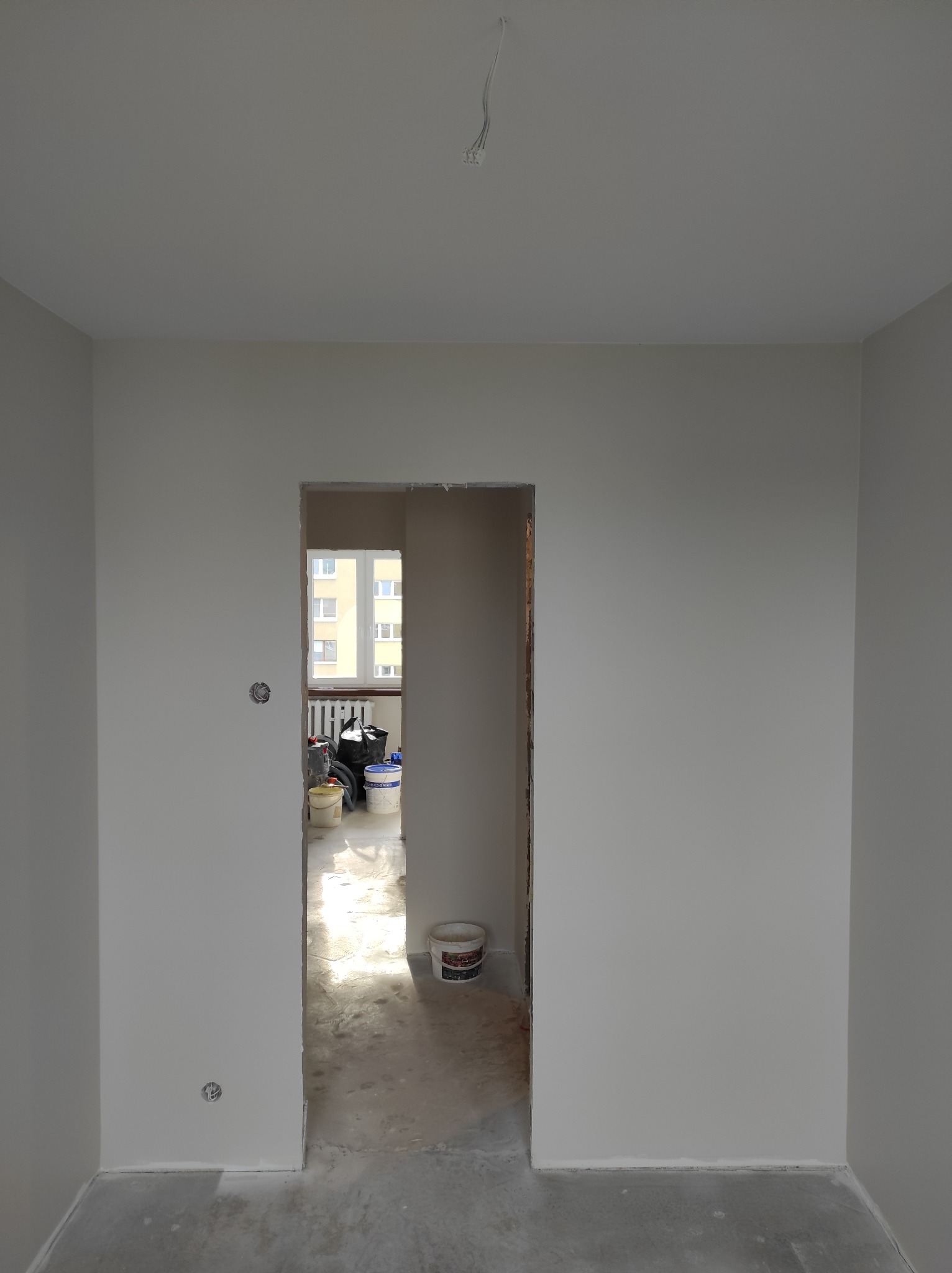 Pomalowane na szaro ściany w pokoju, widoczne puszki elektryczne, surowa podłoga i wiaderka z farbą w przejściu do innego pomieszczenia.