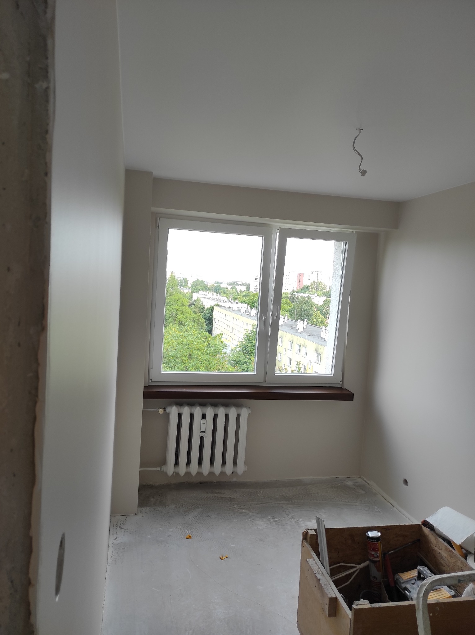 Pokój w trakcie malowania z widocznym oknem, grzejnikiem i narzędziami malarskimi w skrzynce, ściany świeżo pomalowane na jasny kolor, surowy beton na framudze drzwi.