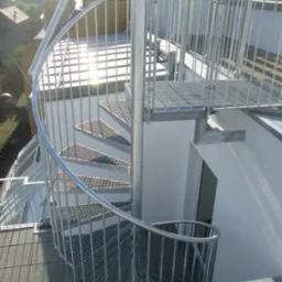 Zewnętrzne schody spiralne z metalu ocynkowanego, prowadzące na dach budynku, z widoczną datą wykonania na dolnym planie.