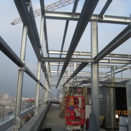 MKO-Konstrukcje SP Z OO - Konstrukcja stalowa budynku w trakcie montażu, widoczny dźwig w tle, miasto w oddali, podnośnik koszowy na pierwszym planie.