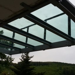 MKO-Konstrukcje SP Z OO - Metalowa konstrukcja z przeszklonym zadaszeniem, widok z dołu na tle zieleni i pochmurnego nieba.