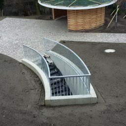 MKO-Konstrukcje SP Z OO - Zewnętrzne schody betonowe z metalową balustradą prowadzące do podziemia, otoczone ziemią przygotowaną pod trawnik, w tle ścieżka z kamiennej kostki i drewniana altana.