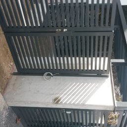 MKO-Konstrukcje SP Z OO - Nowoczesna, grafitowa brama wjazdowa z pionowymi szczeblami, widok z góry, z kablem elektrycznym leżącym na betonowej podstawie.