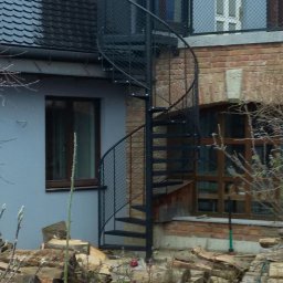 MKO-Konstrukcje SP Z OO - Zewnętrzne metalowe schody spiralne z siatkową balustradą prowadzące na balkon w budynku z cegły i szarego tynku, widoczne składowane drewno opałowe.