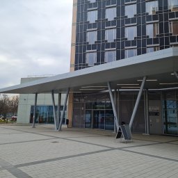 MKO-Konstrukcje SP Z OO - Zadaszone wejście do nowoczesnego budynku z przeszkloną fasadą i ukośnymi metalowymi podporami, widok z zewnątrz na plac wyłożony kostką.