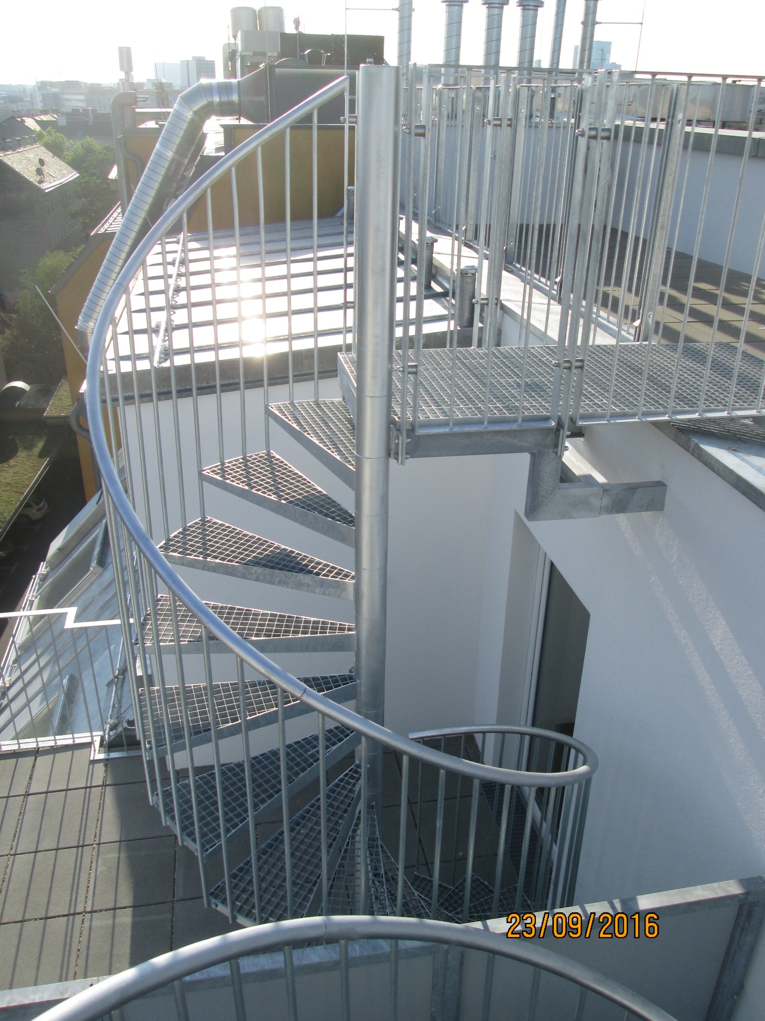 Zewnętrzne schody spiralne z metalu ocynkowanego, prowadzące na dach budynku, z widoczną datą wykonania na dolnym planie.