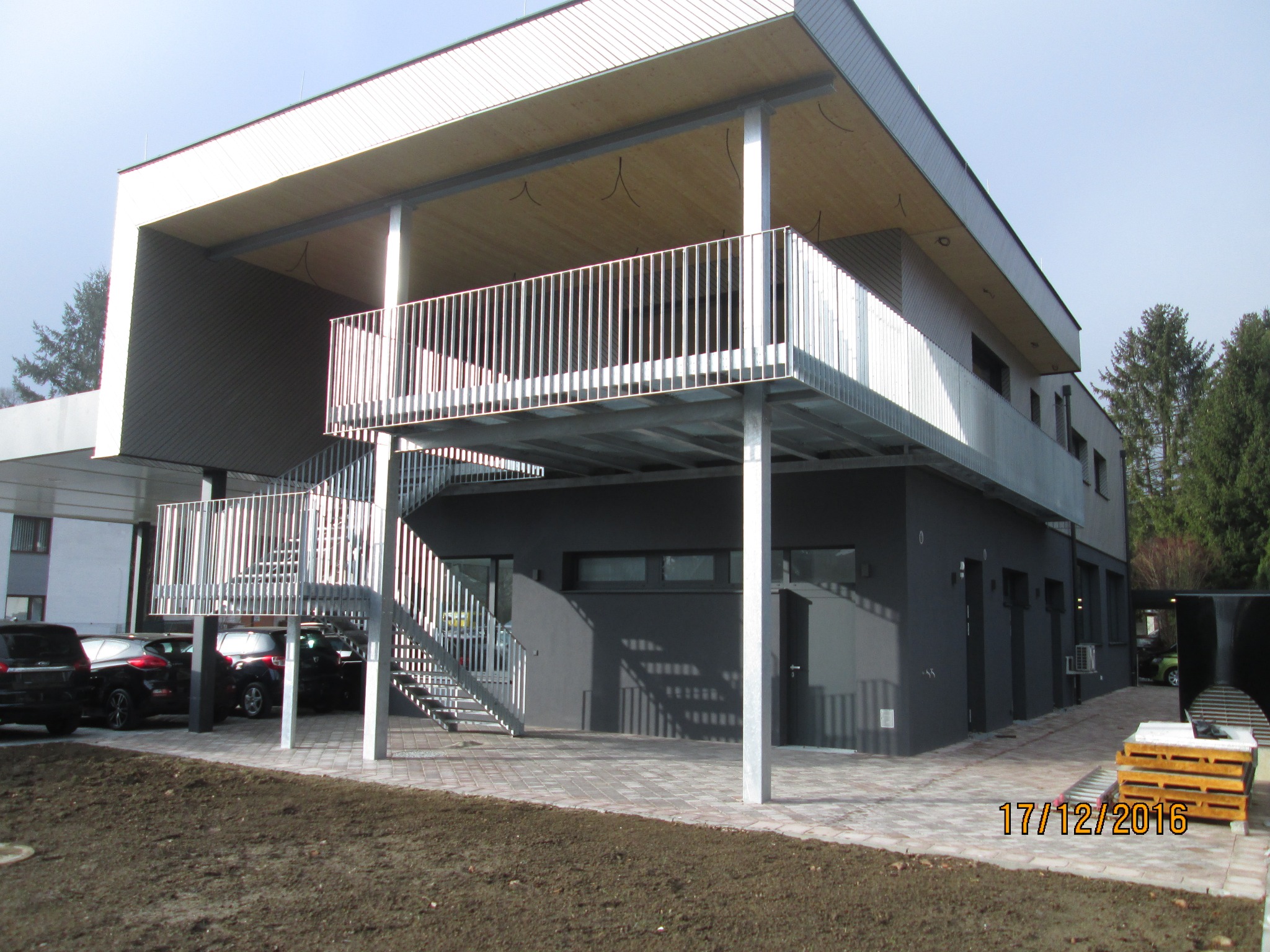 Nowoczesny budynek z zewnętrznymi schodami i balkonem z metalowymi barierkami, widoczne zaparkowane samochody i teren przygotowany pod nasadzenia.