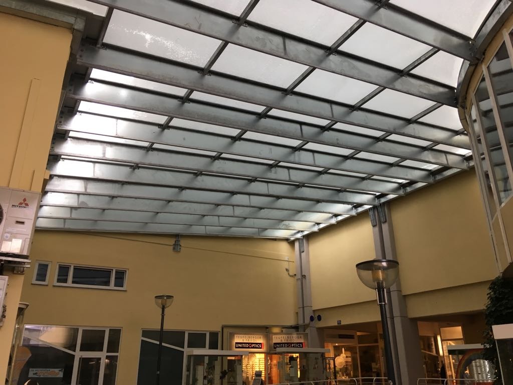 Metalowa konstrukcja z przezroczystymi panelami tworząca zadaszenie pasażu handlowego, widoczna z dołu, z elementami oświetlenia i klimatyzacji.