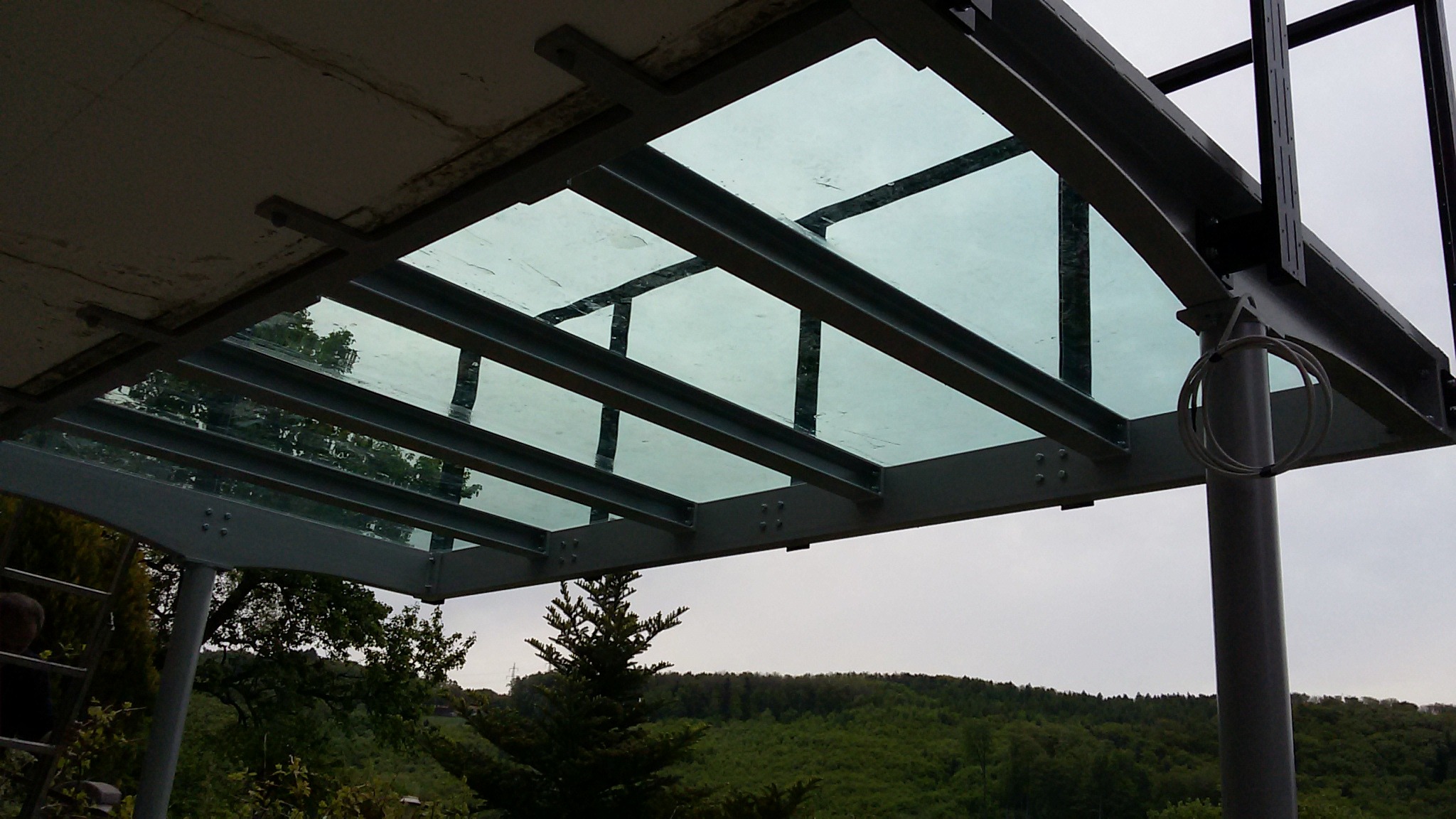 Metalowa konstrukcja z przeszklonym zadaszeniem, widok z dołu na tle zieleni i pochmurnego nieba.