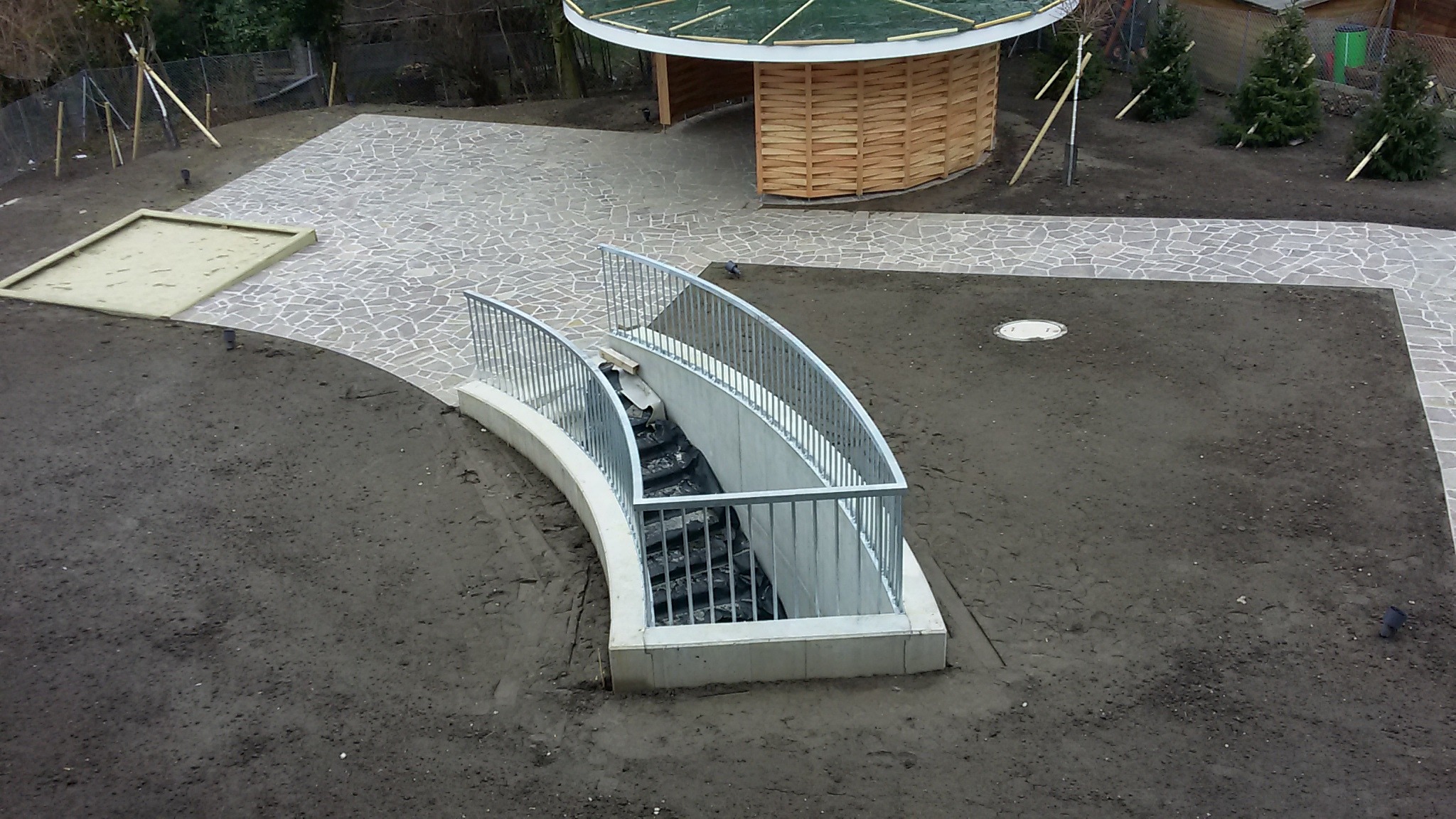 Zewnętrzne schody betonowe z metalową balustradą prowadzące do podziemia, otoczone ziemią przygotowaną pod trawnik, w tle ścieżka z kamiennej kostki i drewniana altana.