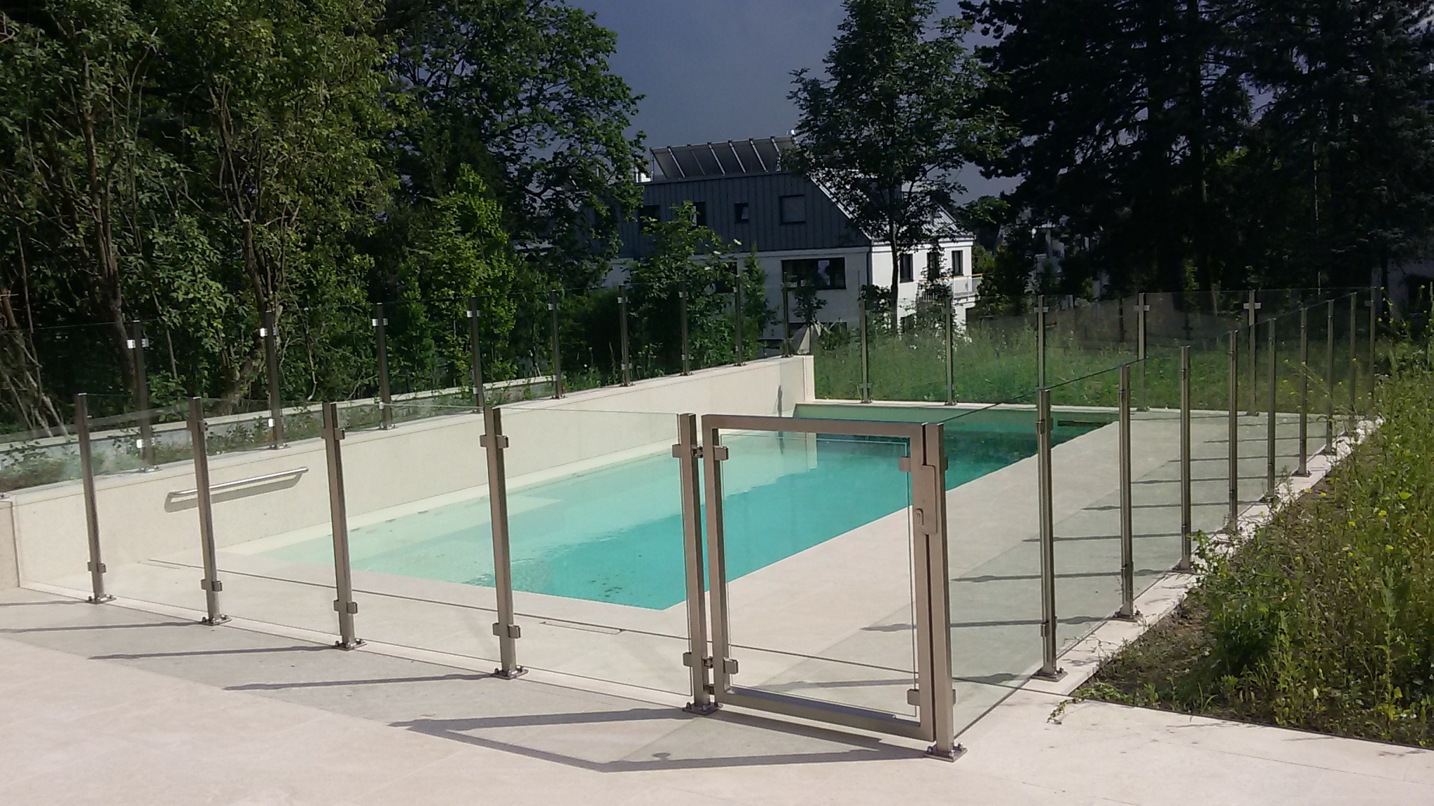Szklana balustrada z metalowymi słupkami otaczająca basen zewnętrzny z otwartą furtką, widok na dom z ciemnym dachem i zieleń w tle.