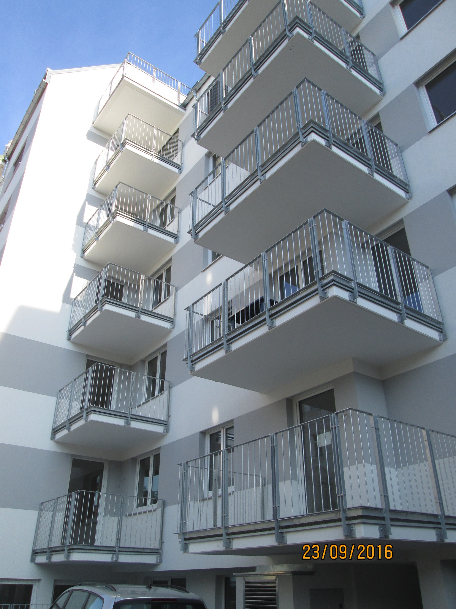 Wielopiętrowy budynek z balkonami zabezpieczonymi metalowymi balustradami o prostej, pionowej konstrukcji, widok z dołu na tle błękitnego nieba.