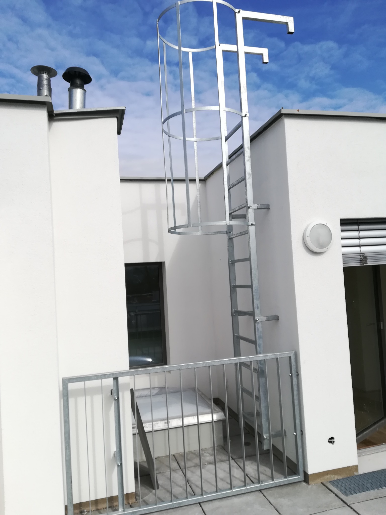 Zewnętrzna metalowa drabina ewakuacyjna z zabezpieczeniem klatkowym, zamontowana na białym budynku z balkonem i metalową balustradą, widok na niebieskie niebo.