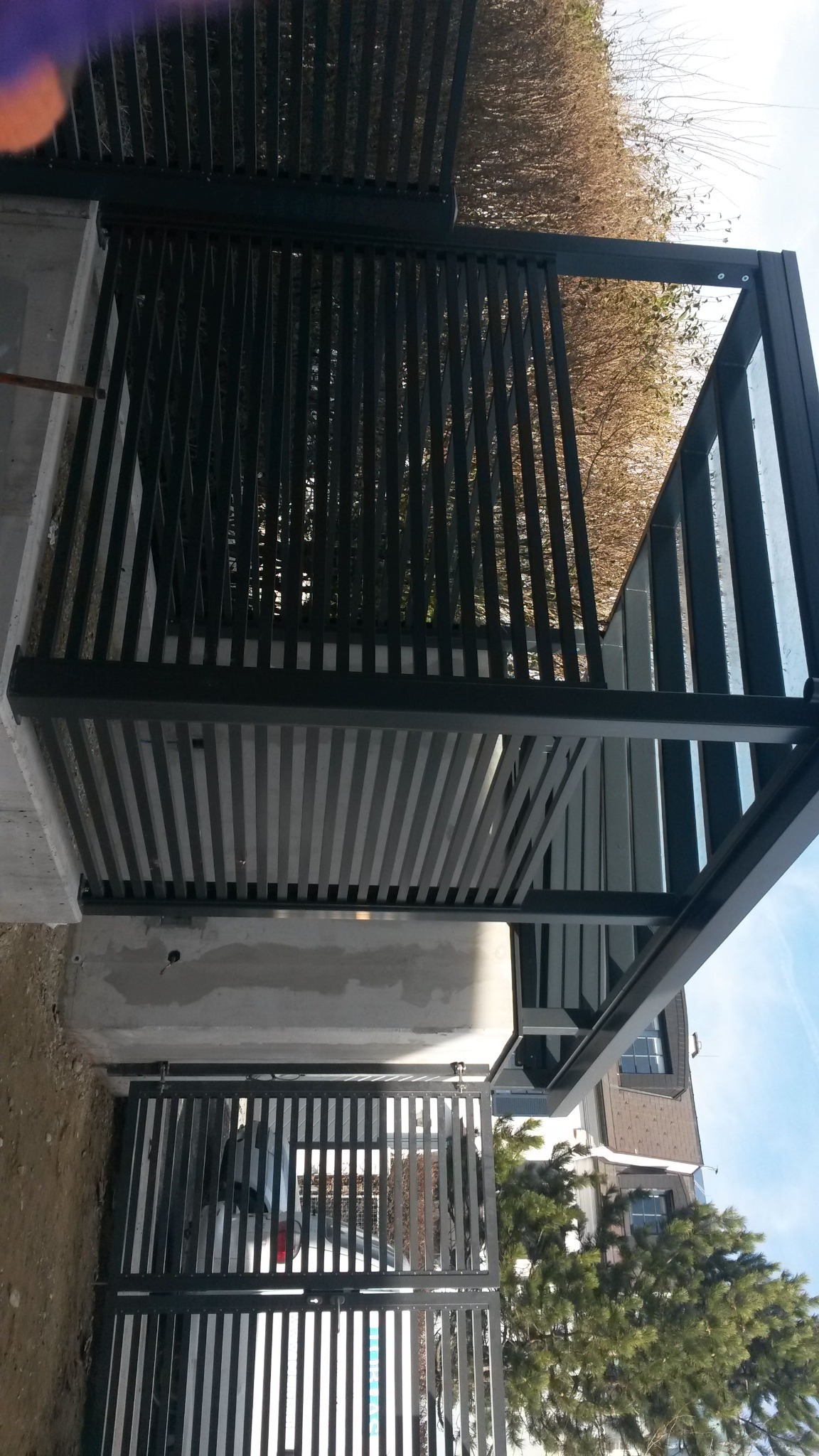 Nowoczesna, metalowa konstrukcja z pionowymi listwami, prawdopodobnie brama lub ogrodzenie, z widocznym betonowym fundamentem i fragmentem budynku w tle, sfotografowana pod kątem, ukazująca detale...