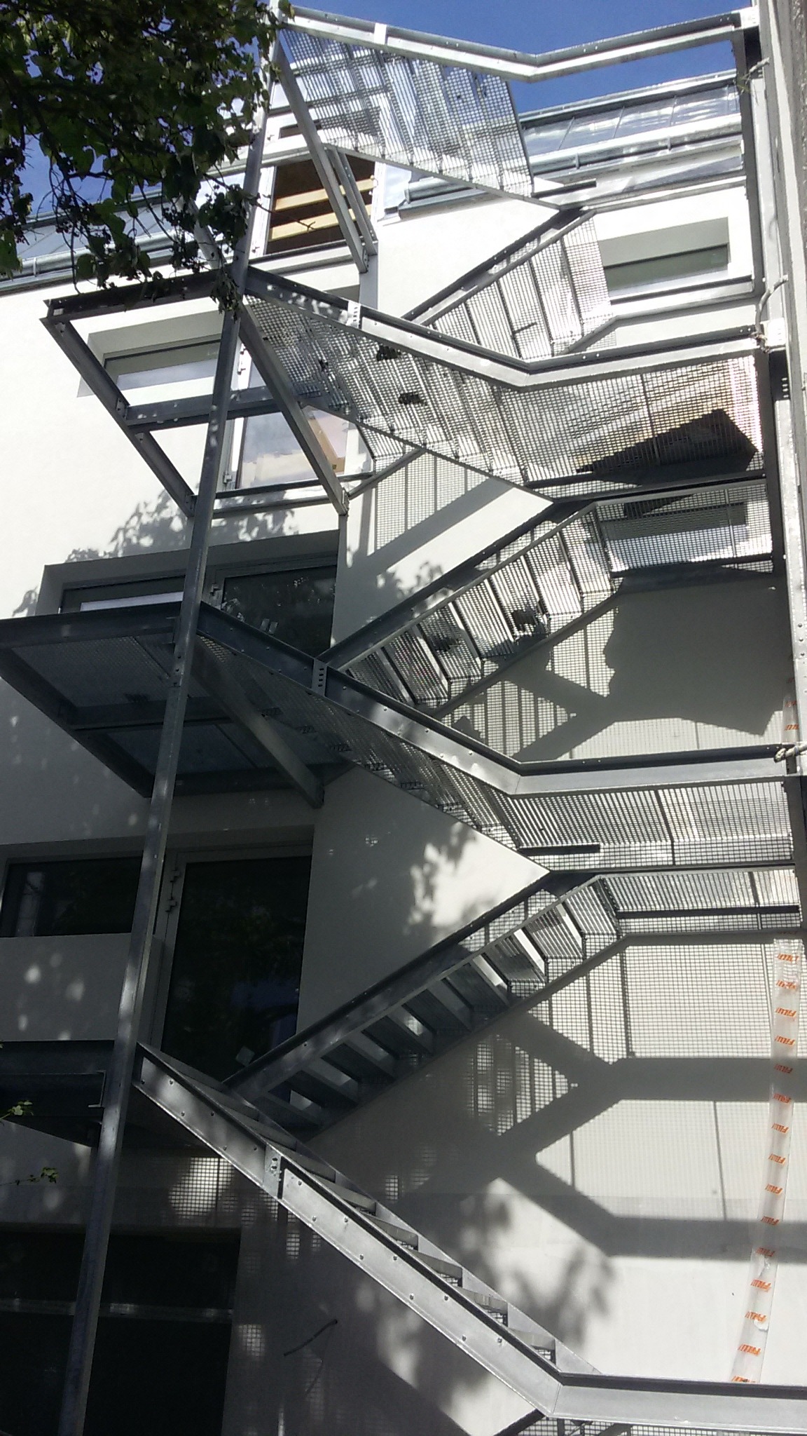 Zewnętrzne schody ewakuacyjne z metalową konstrukcją i ażurowymi stopniami, zamontowane na elewacji budynku z widocznymi oknami i fragmentami zieleni.