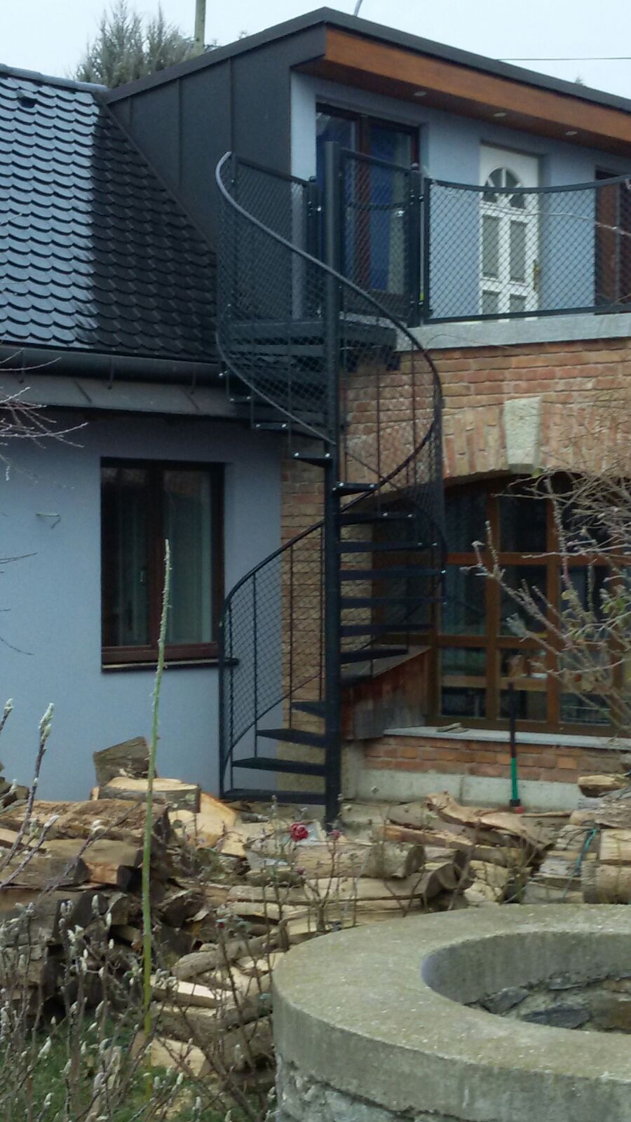 Zewnętrzne metalowe schody spiralne z siatkową balustradą prowadzące na balkon w budynku z cegły i szarego tynku, widoczne składowane drewno opałowe.