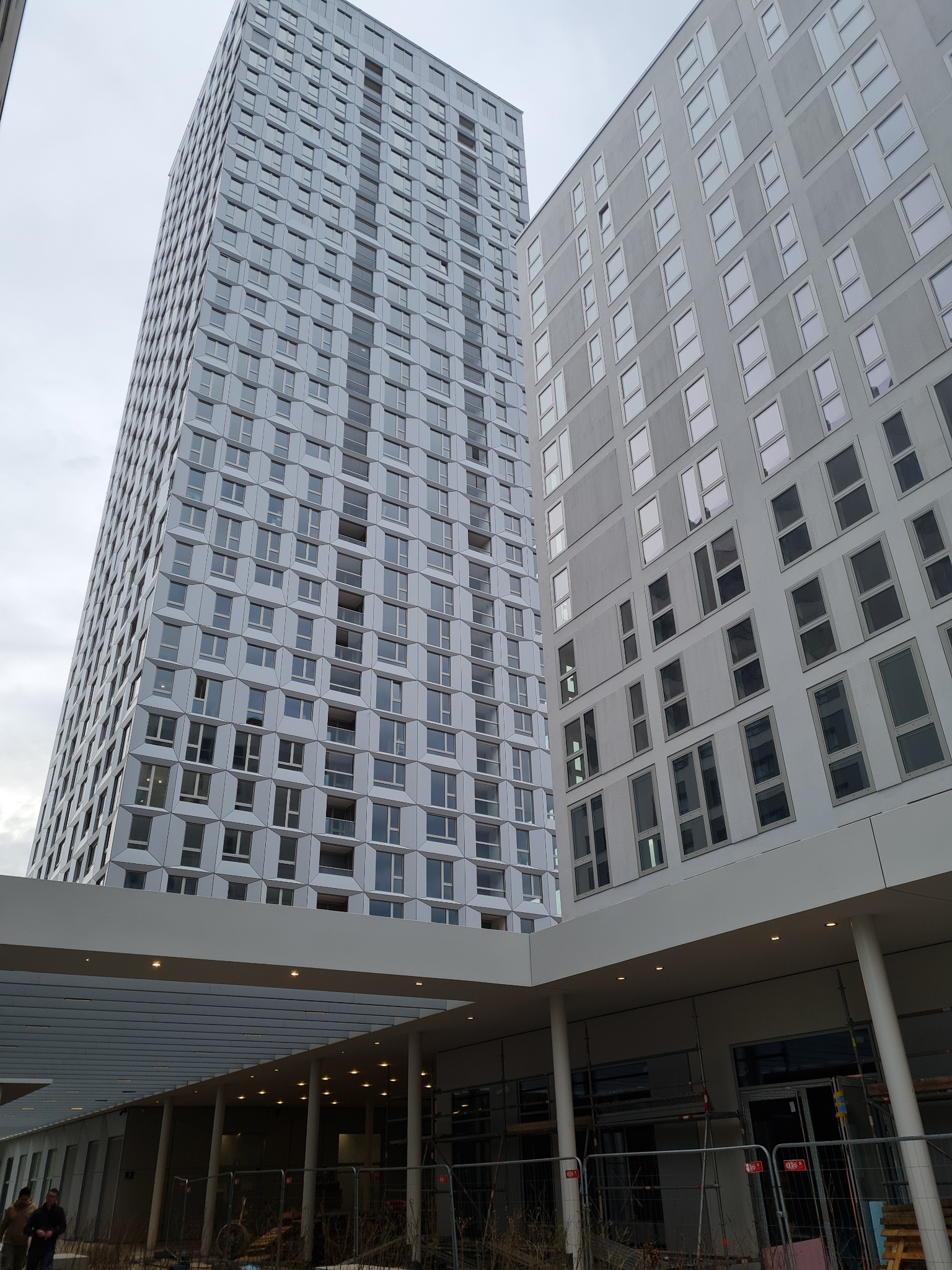 Dwa nowoczesne, wysokie budynki w Nowym Targu, jeden z elewacją w geometryczne wzory, drugi z prostymi oknami, widziane z dołu spod zadaszenia w trakcie budowy.