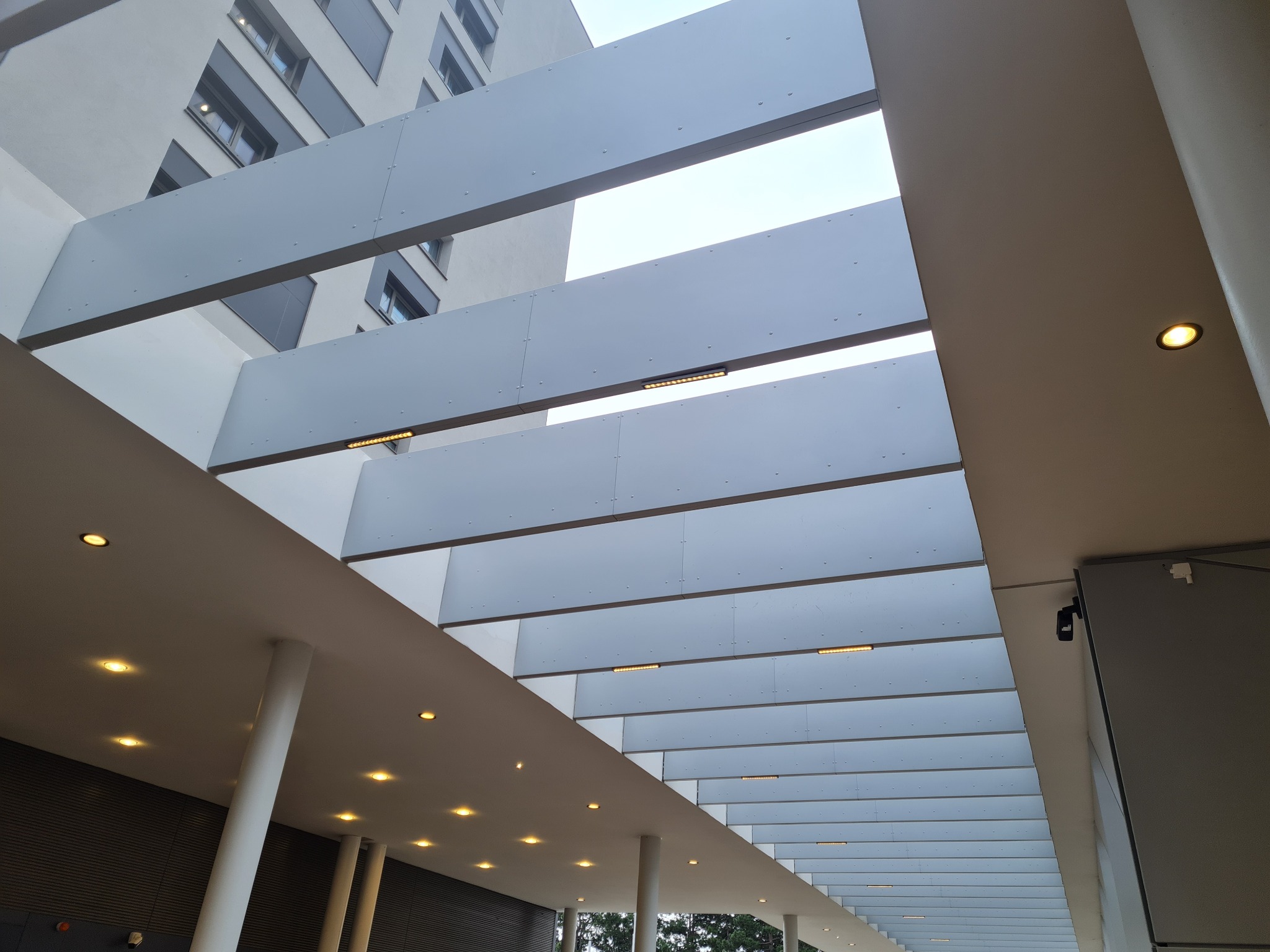 Nowoczesna konstrukcja z metalowych paneli tworząca zadaszenie nad przestrzenią publiczną, widok z dołu na regularny układ paneli z wbudowanym oświetleniem LED, budynek w tle.