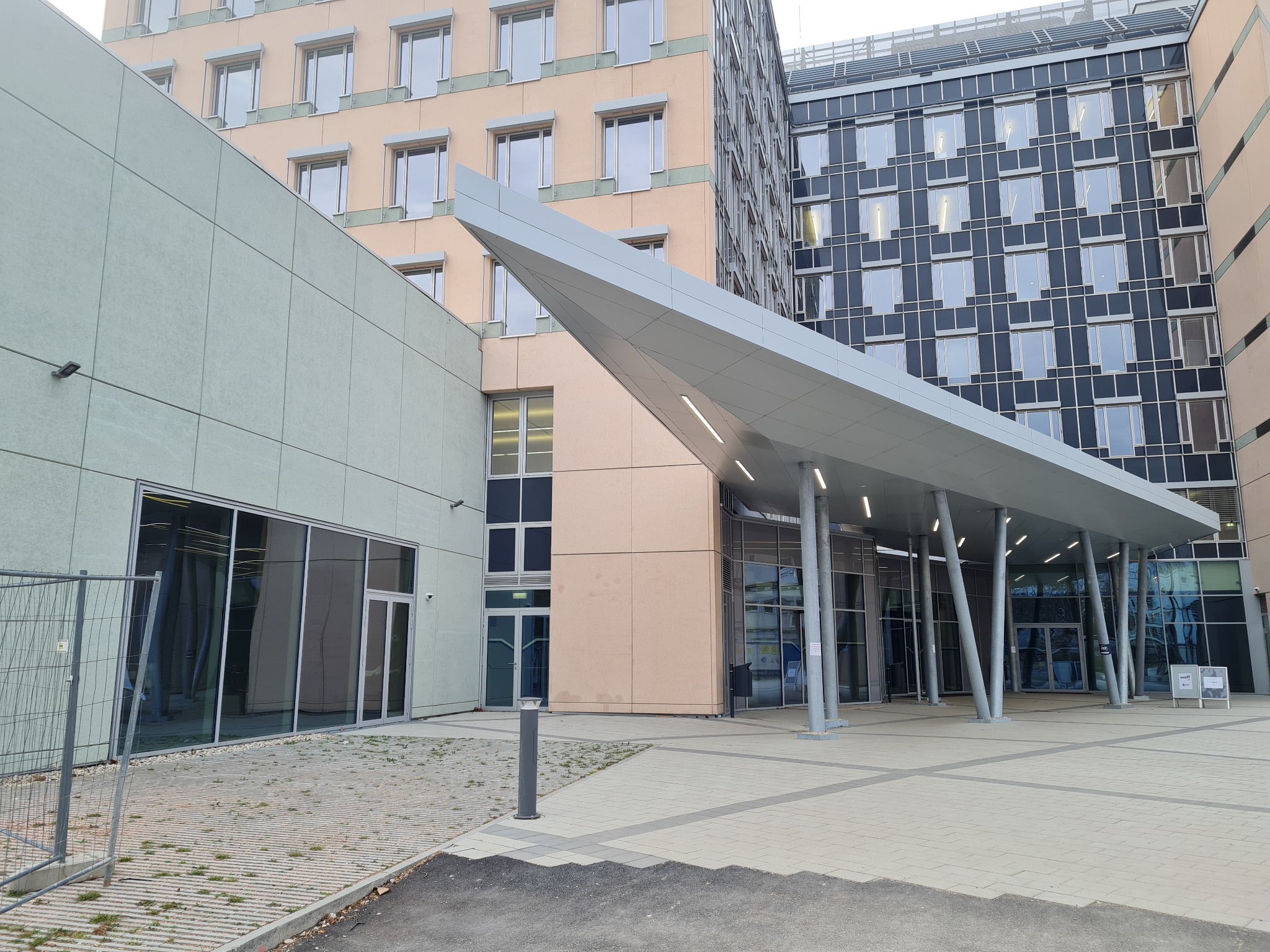 Nowoczesne wejście do budynku biurowego z zadaszeniem wspartym na ukośnych kolumnach, widok z zewnątrz na szare płytki chodnikowe i trzy budynki w tle o różnej architekturze.