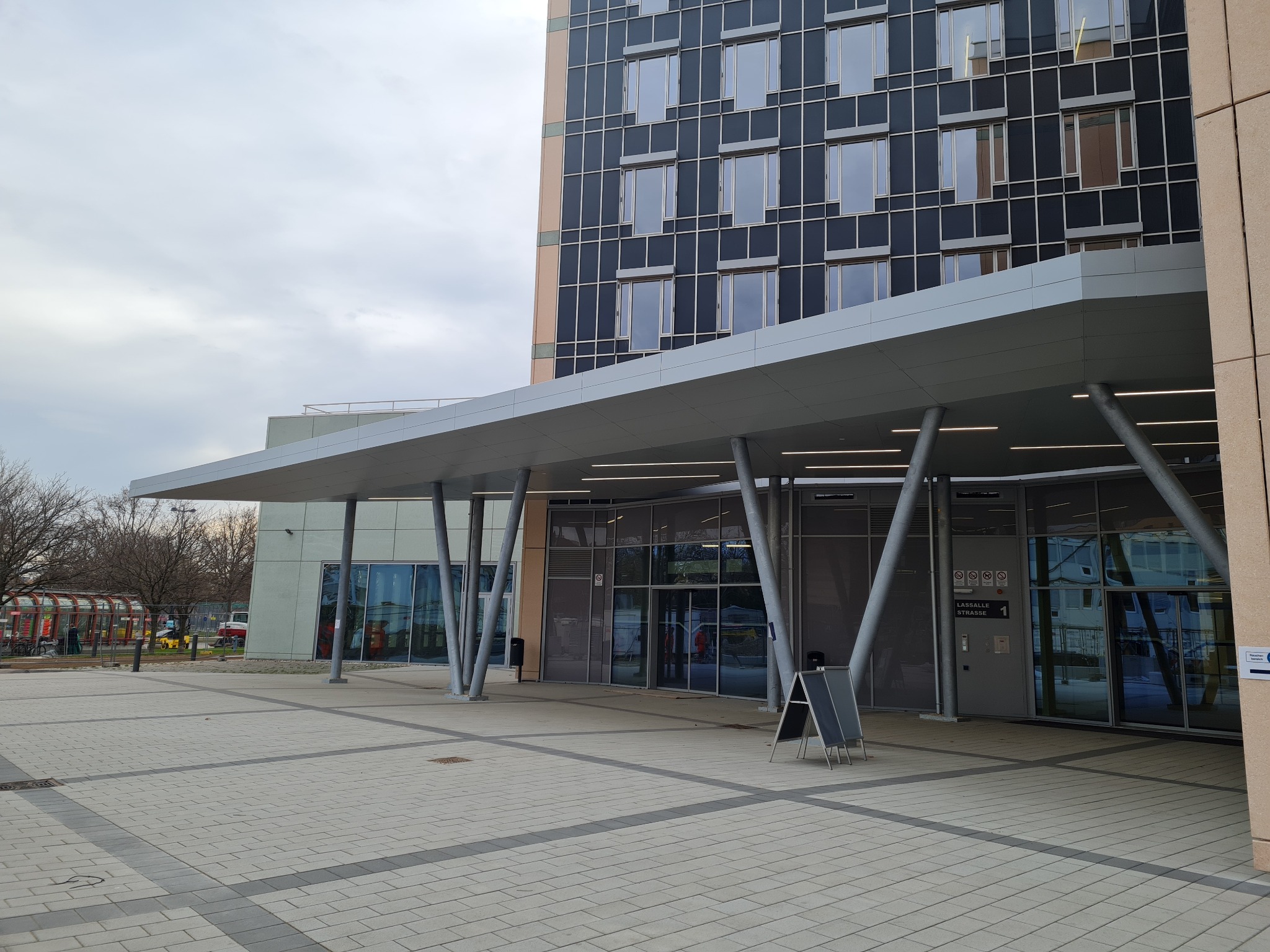 Zadaszone wejście do nowoczesnego budynku z przeszkloną fasadą i ukośnymi metalowymi podporami, widok z zewnątrz na plac wyłożony kostką.