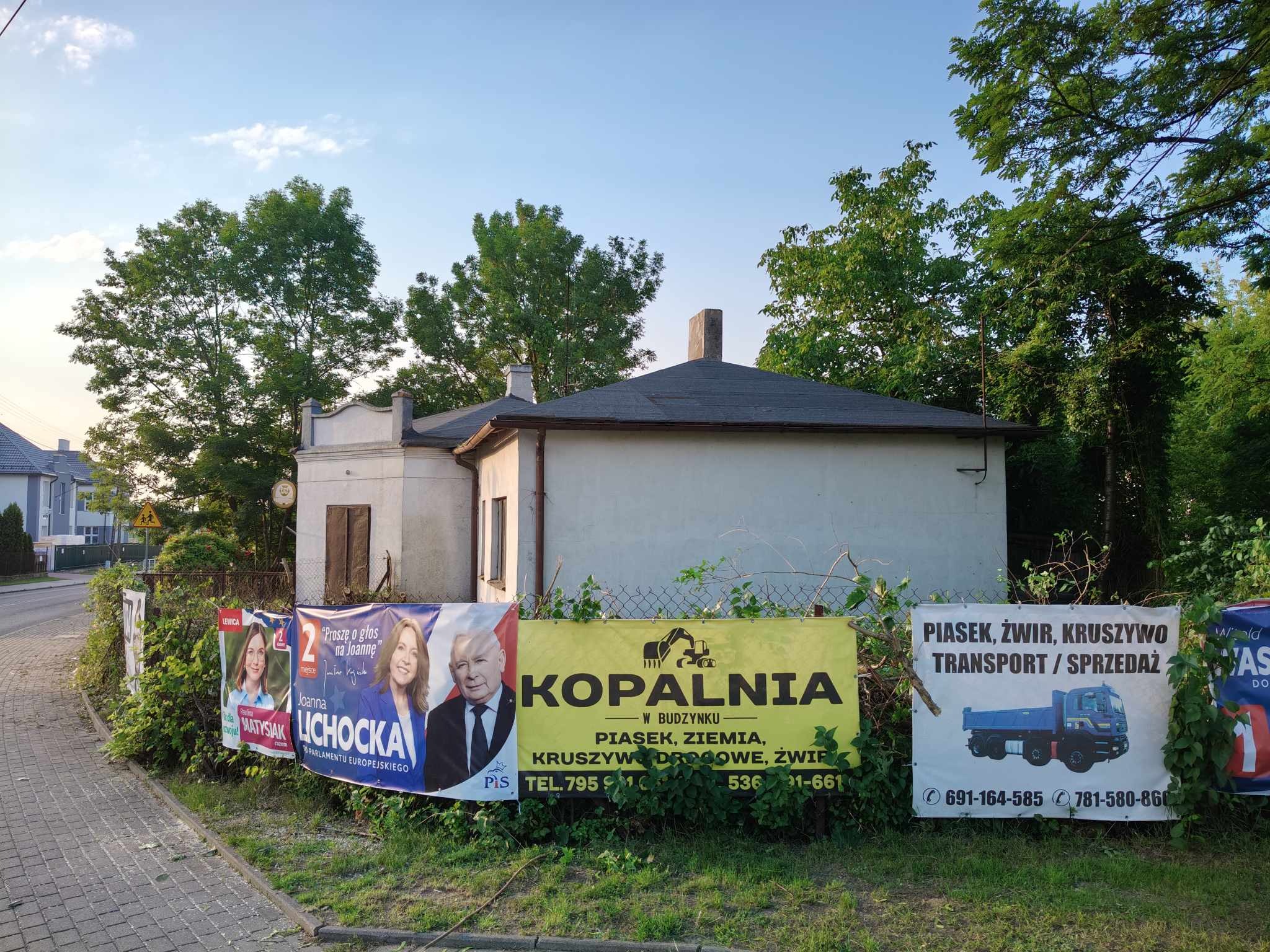 Ogrodzenie posesji w Zgierzu z reklamami usług związanych z kopalinami i transportem kruszyw, na tle budynku i drzew.