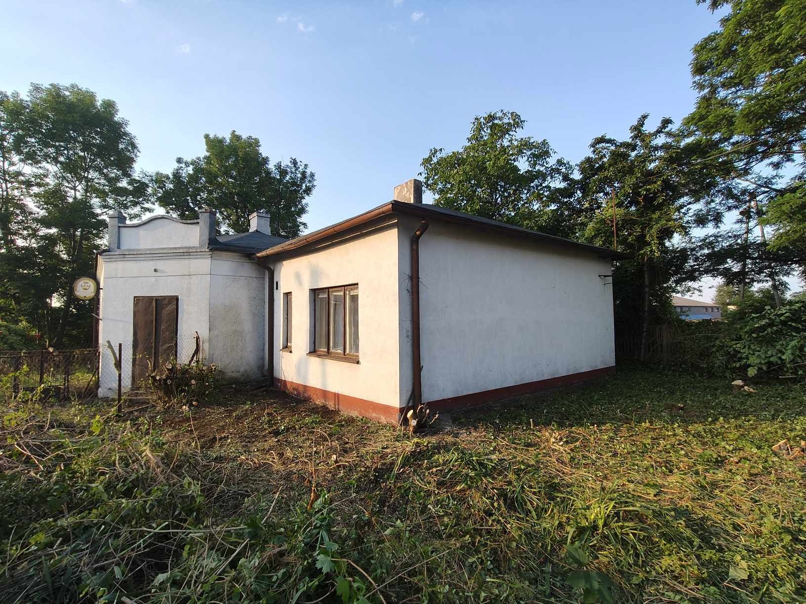 Kępy ściętych gałęzi i krzewów przed białym budynkiem z dobudówką, widoczne drzewa w tle.