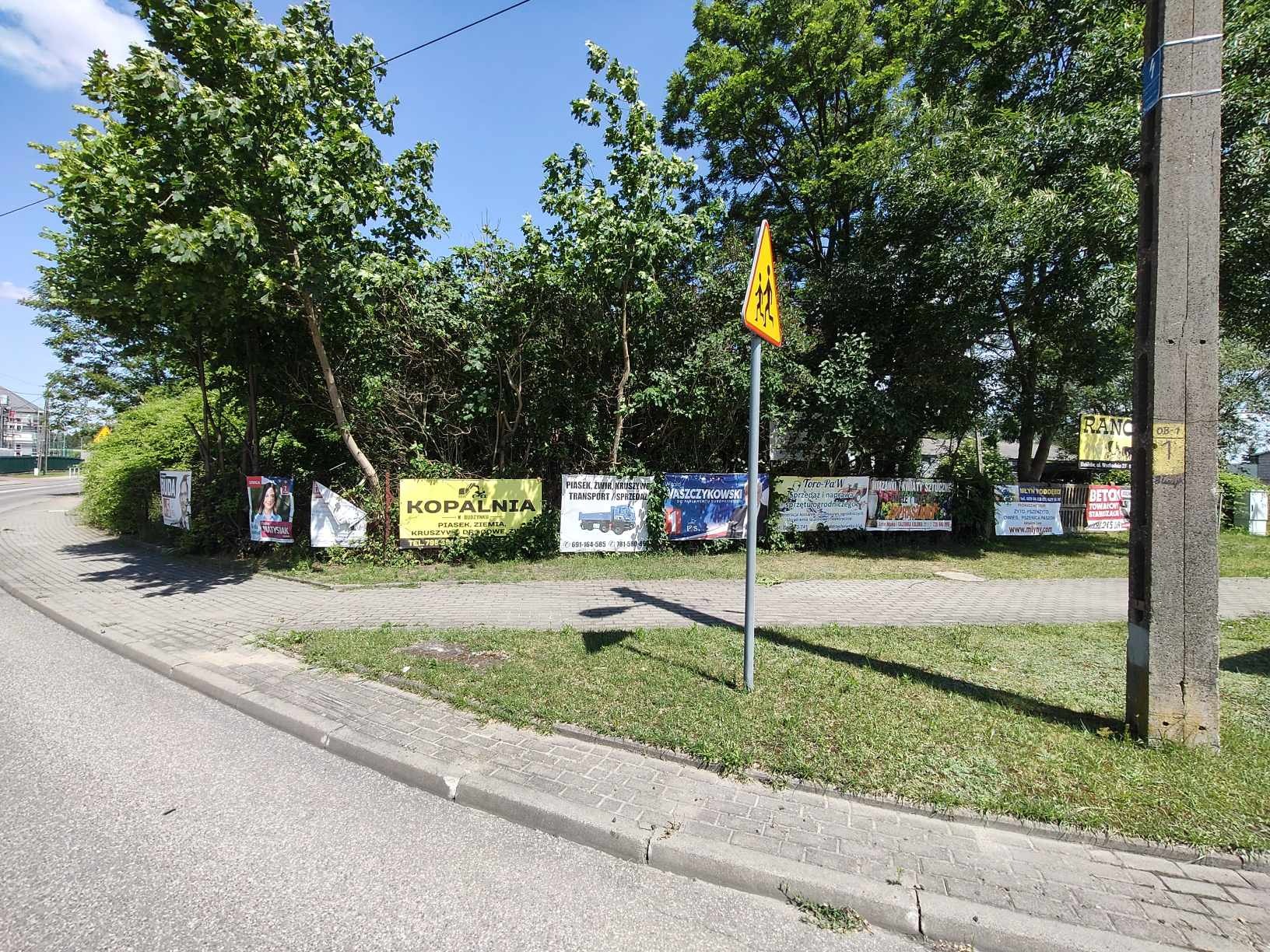 Tablice reklamowe różnych firm (kopalnia, transport, kwiaty, beton) umieszczone na tle drzew i krzewów przy chodniku i ulicy, w tle znak drogowy ostrzegający o przejściu dla pieszych.