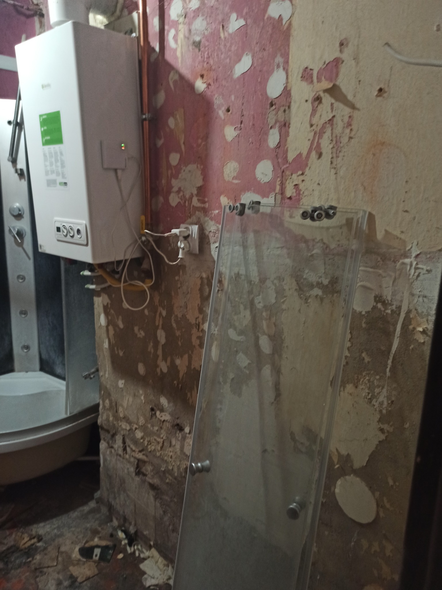 Demontaż kabiny prysznicowej w łazience ze ścianami w trakcie przygotowania do malowania, widoczny wiszący piec gazowy i wystające rury.