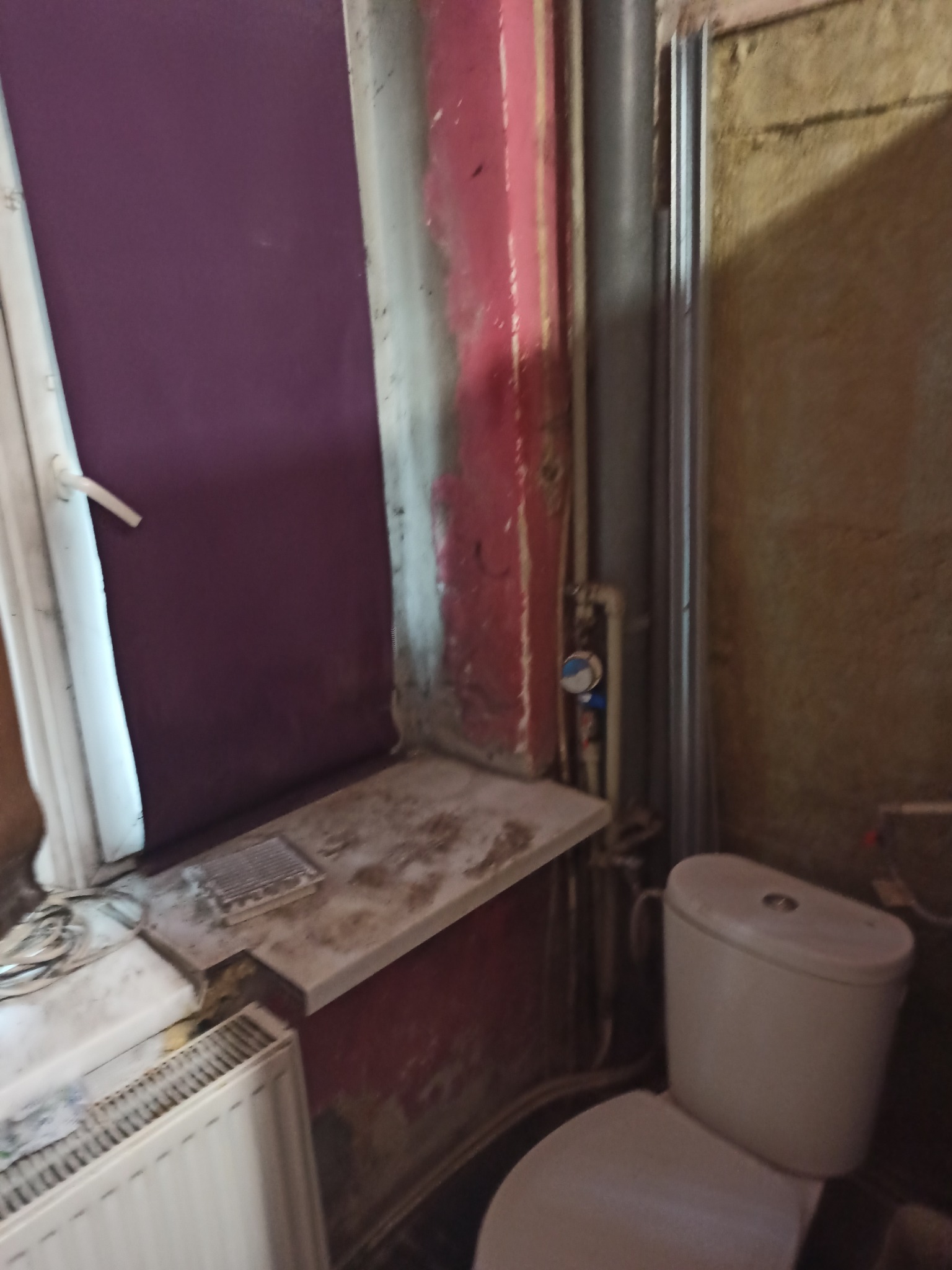 Łazienka w trakcie remontu: widoczna toaleta, grzejnik, okno z fioletową ścianą, ślady starej farby i rury.