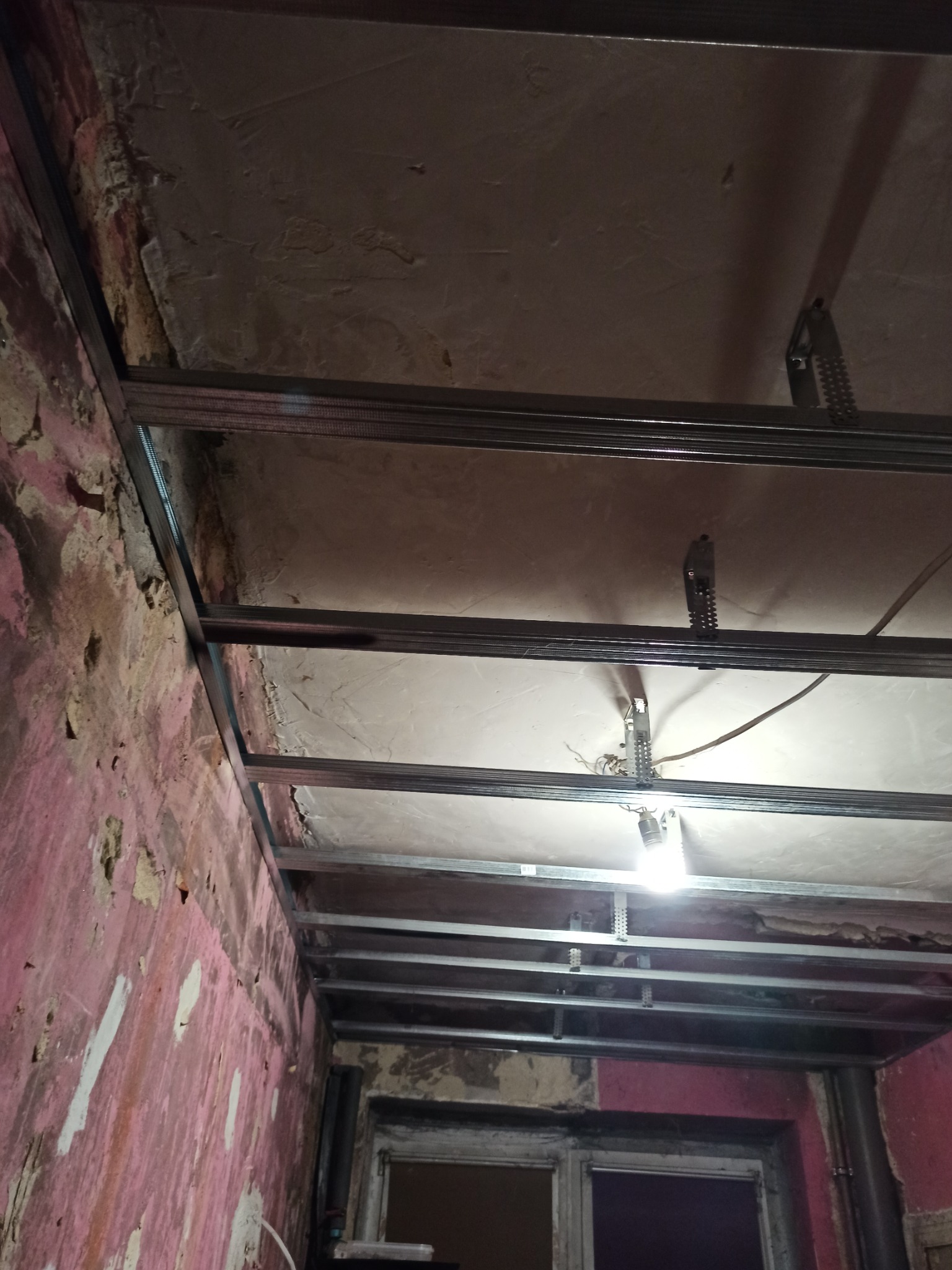 Metalowy stelaż sufitu podwieszanego z widocznymi elementami montażowymi, w tle fragment ściany z resztkami starej farby i okno.