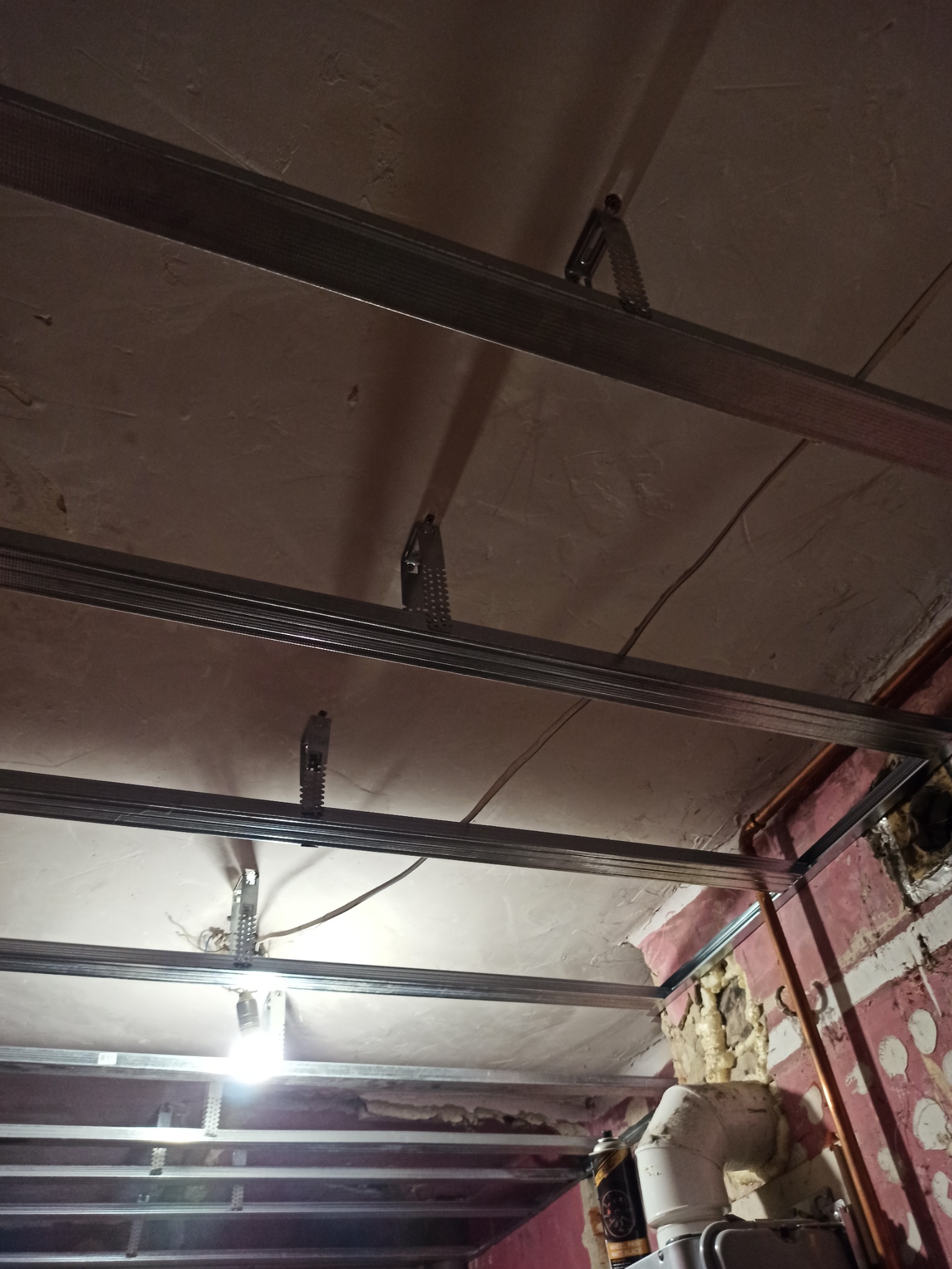 Metalowa konstrukcja sufitu podwieszanego w trakcie montażu, widoczne profile, zawiesia i instalacja elektryczna. Ściana z widocznymi śladami prac remontowych w tle.