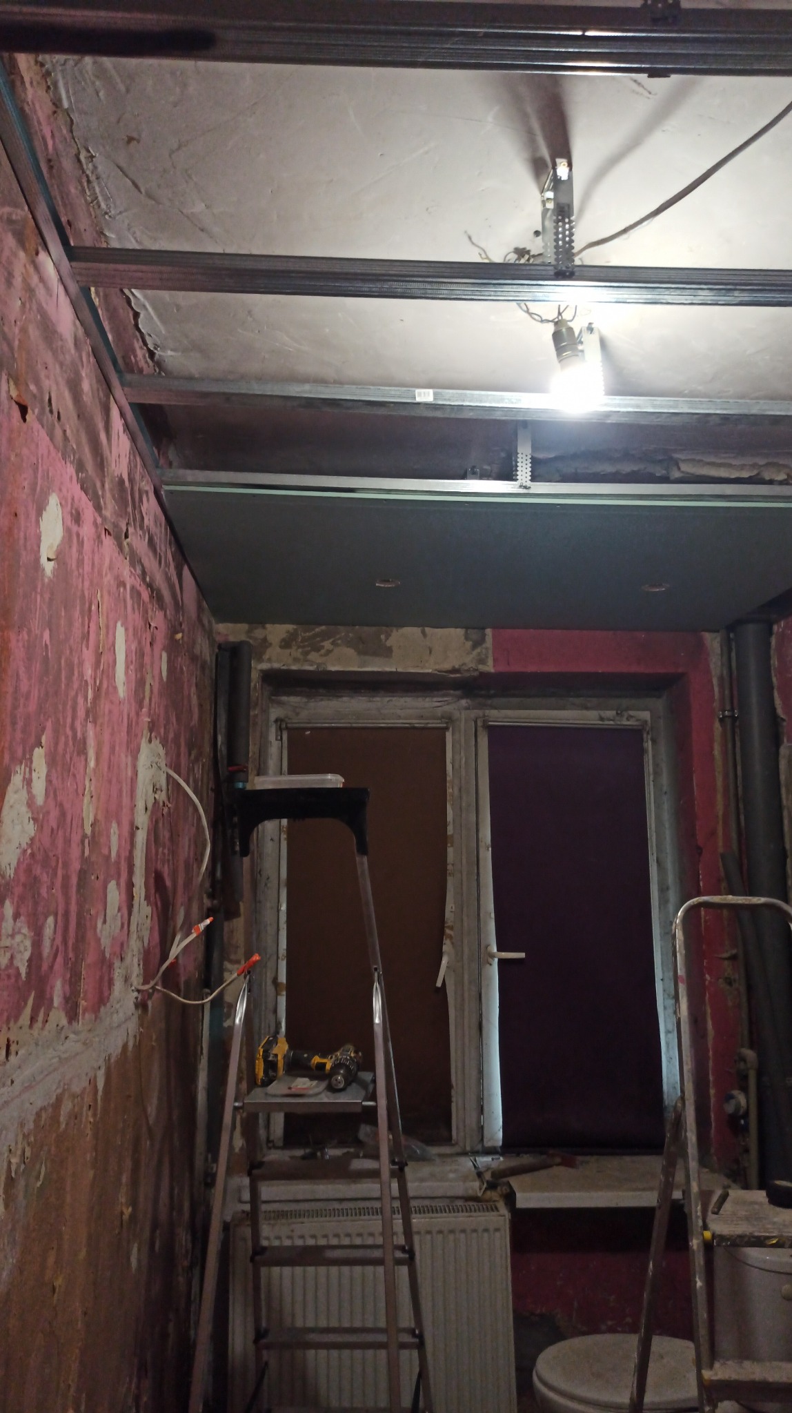 Remont łazienki w trakcie: widoczne metalowe profile sufitowe, częściowo położona płyta gipsowa, drabina z narzędziami i fragmenty starej instalacji sanitarnej.