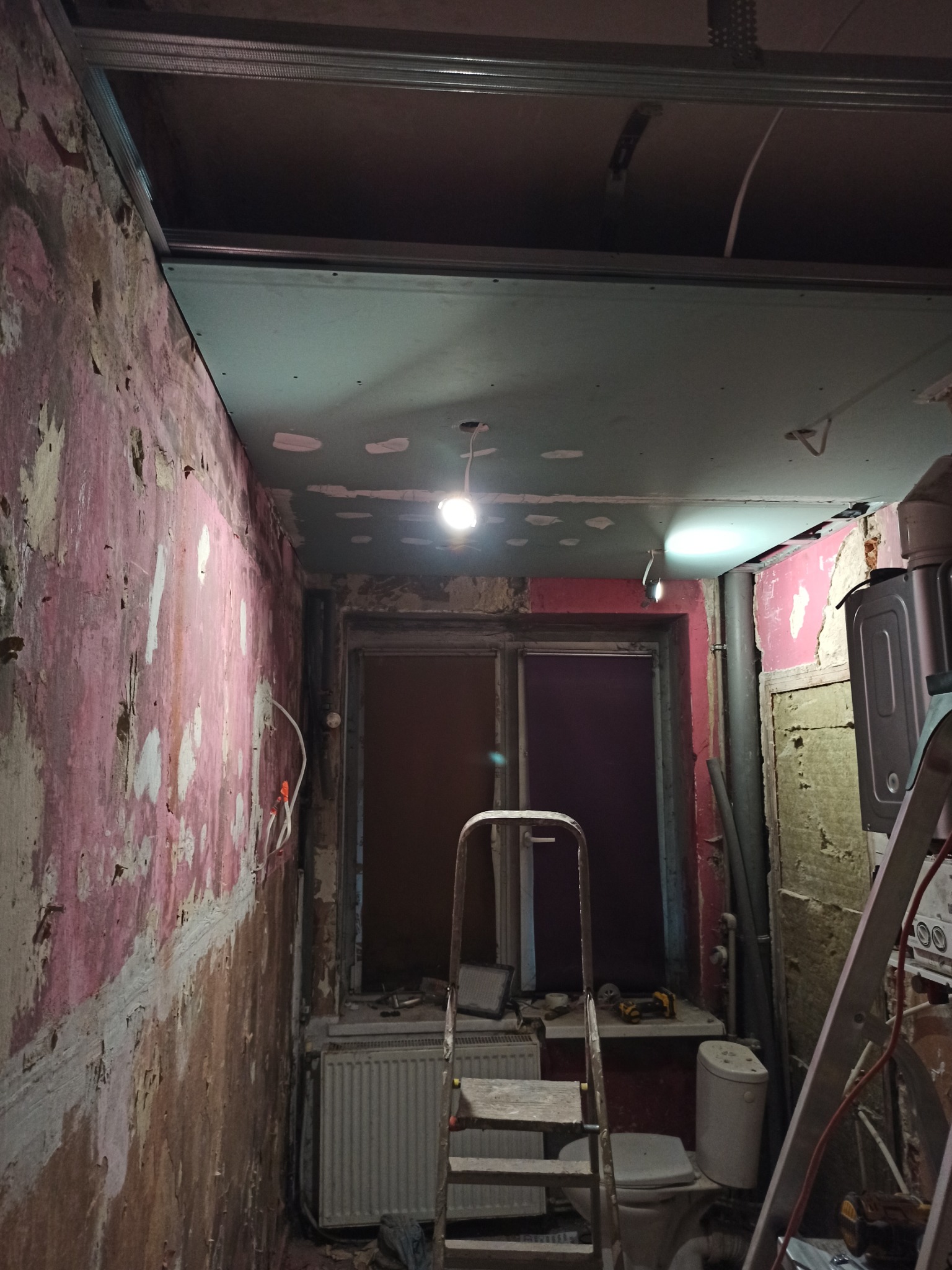 Łazienka w trakcie remontu: ściany ze śladami starej farby, częściowo zamontowany sufit podwieszany, drabina, grzejnik i toaleta.